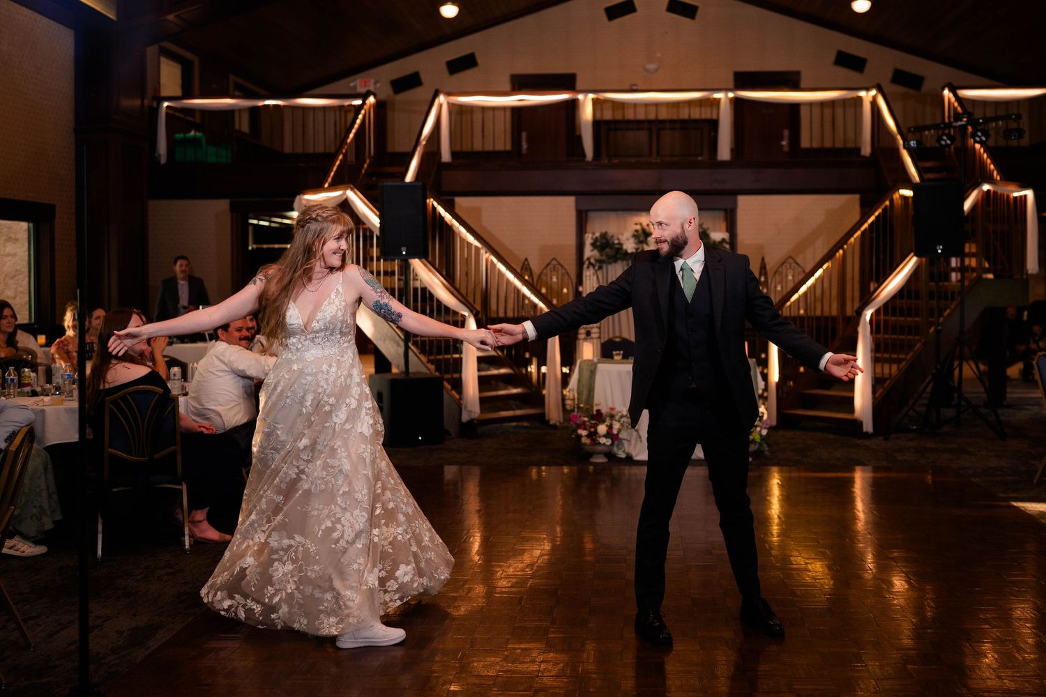 Bride in lace gown and bald groom sharing first dance on polished wooden dance floor.