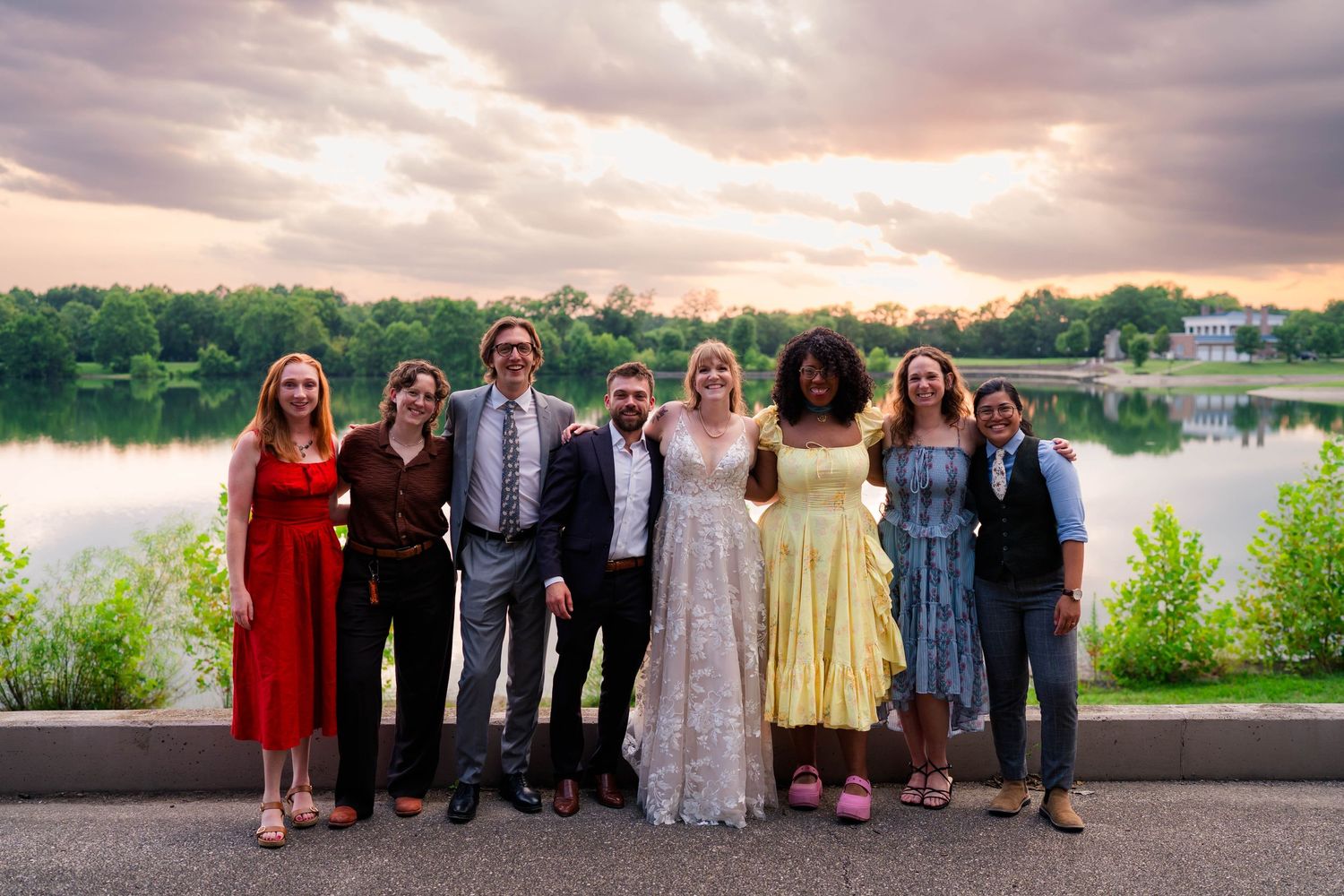 Wedding party poses for a group photo at sunset beside a serene lake with dramatic clouds.