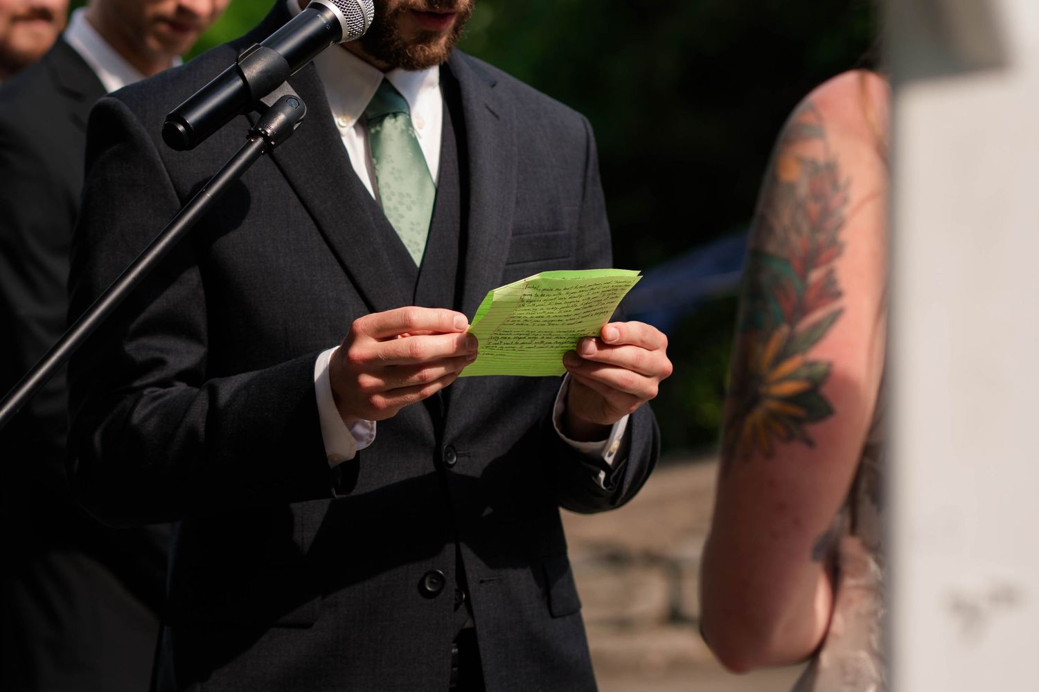 Groom reads heartfelt vows from a green card at outdoor wedding ceremony microphone.