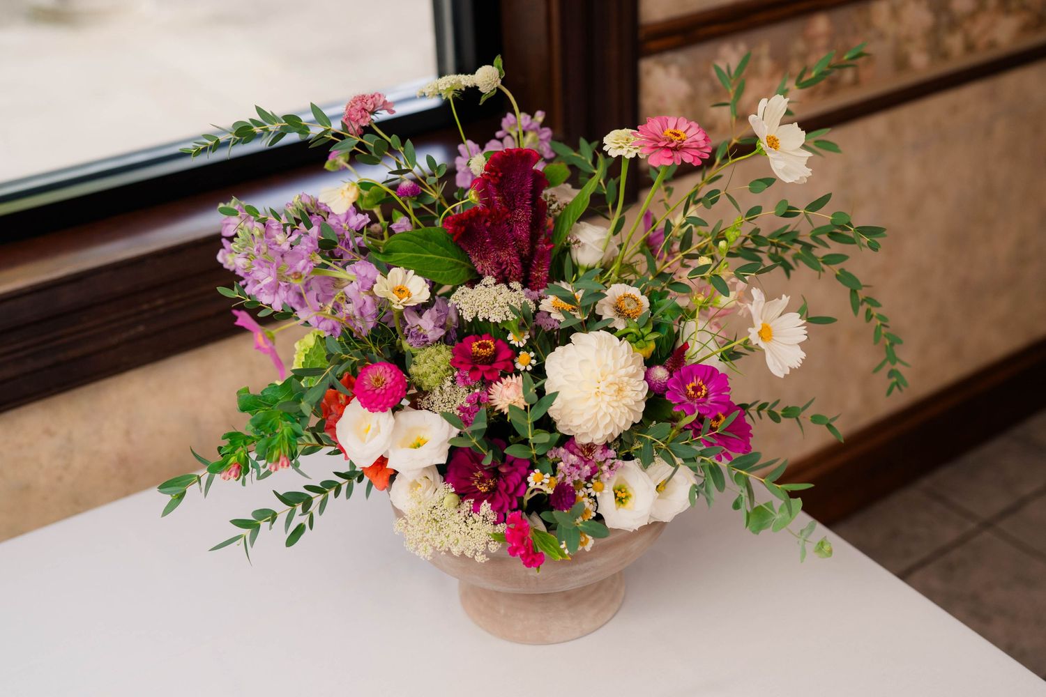 Lush floral arrangement in a beige urn with pink, white, and burgundy blooms and greenery on a white table.