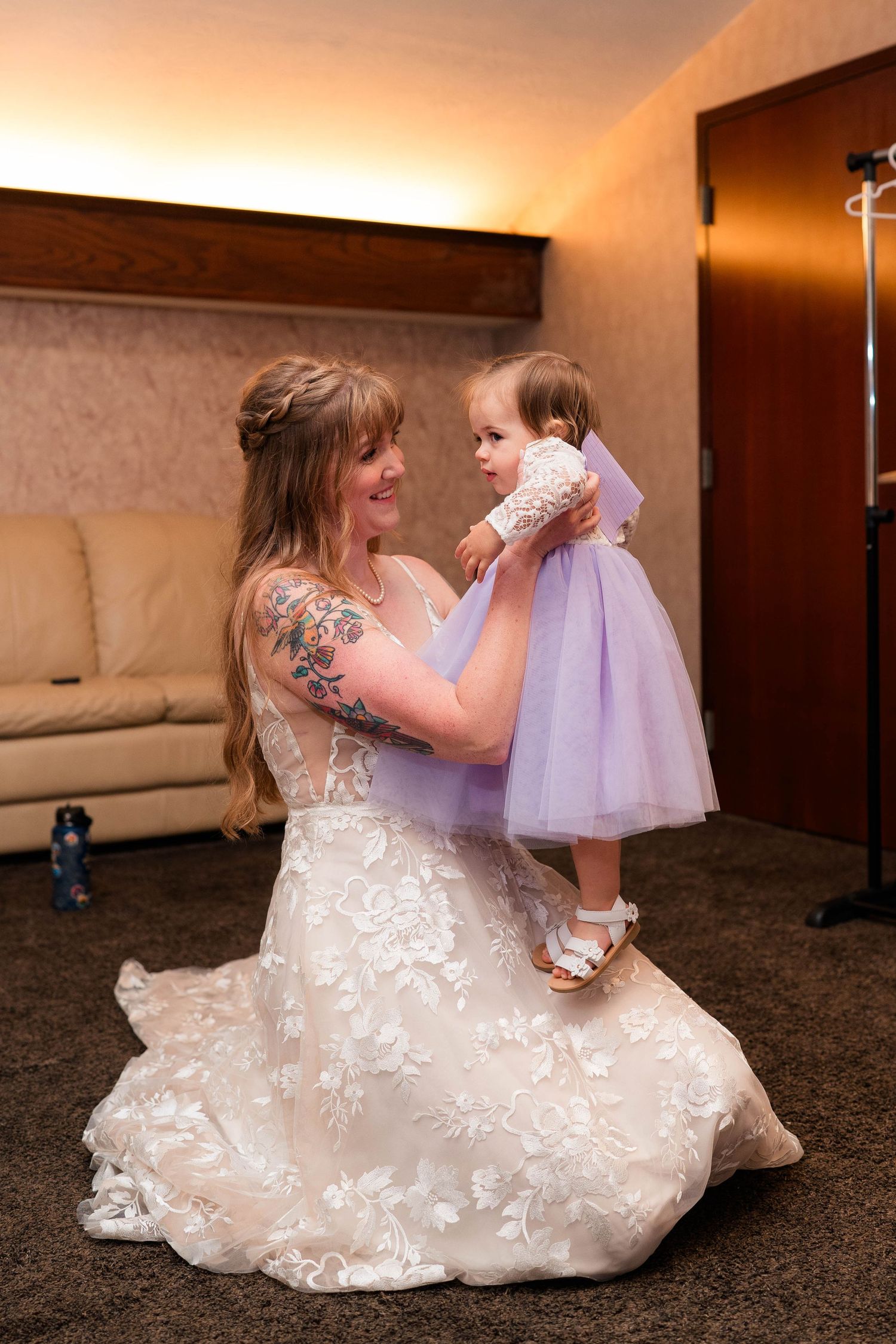 Bride in lace gown holding a toddler girl in a lavender tulle dress indoors.
