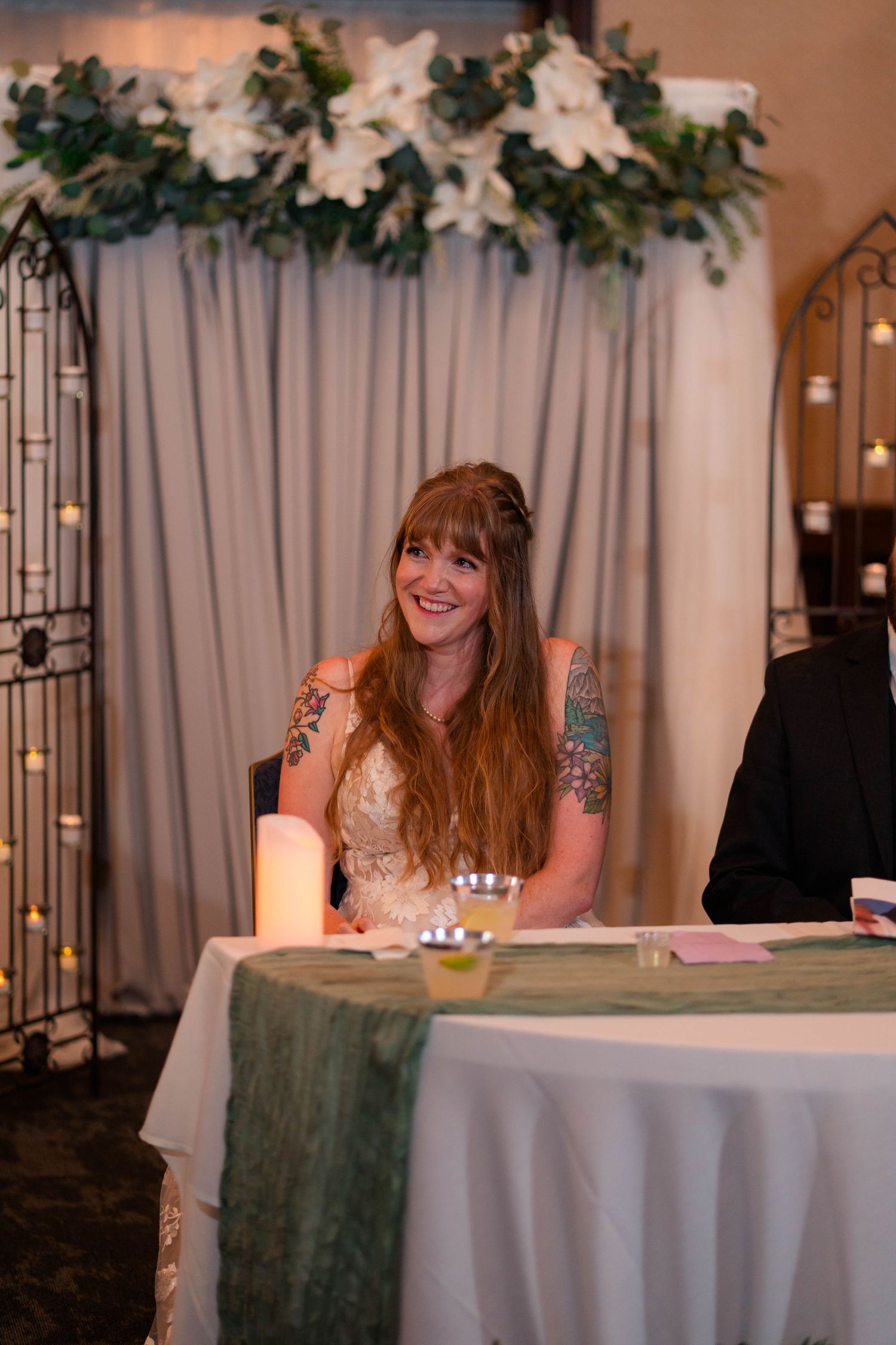 Smiling tattooed bride in lace gown seated at sweetheart table adorned with white floral arch.