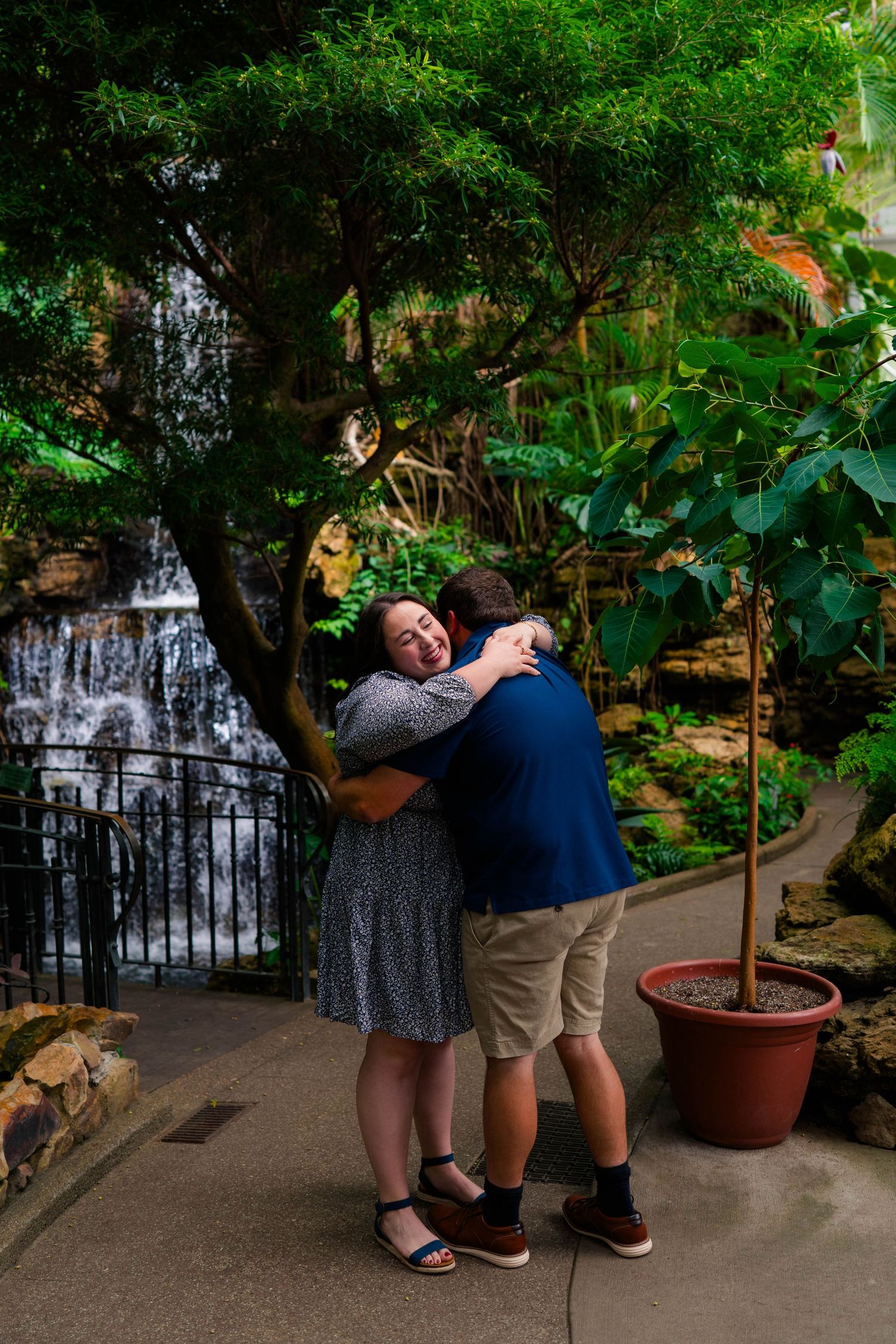 Couple sharing a warm embrace near a waterfall and tropical trees in a botanical garden.