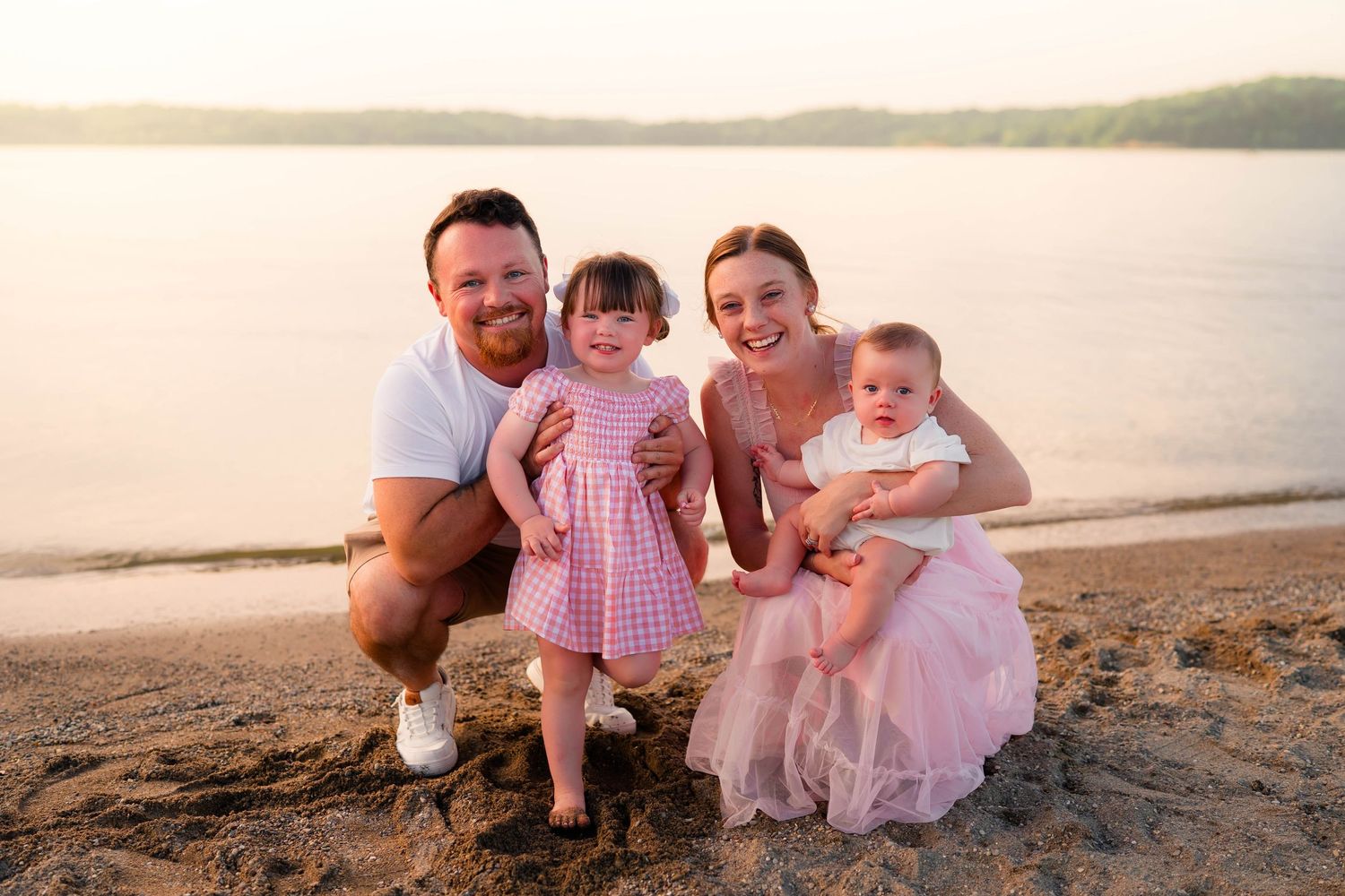 Smiling family of four posing on a sandy beach at golden hour with two young children.