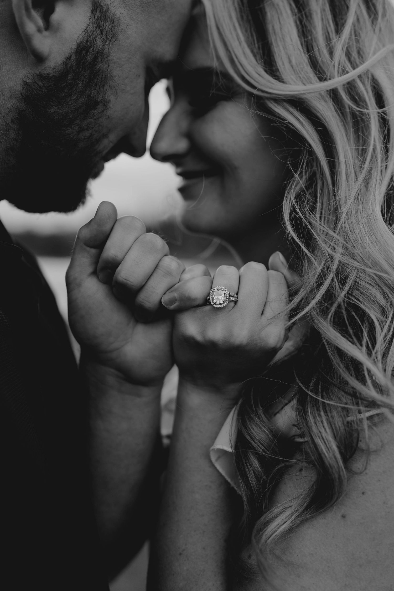 Black and white close-up of an engaged couple touching foreheads, showcasing a stunning halo diamond engagement ring.