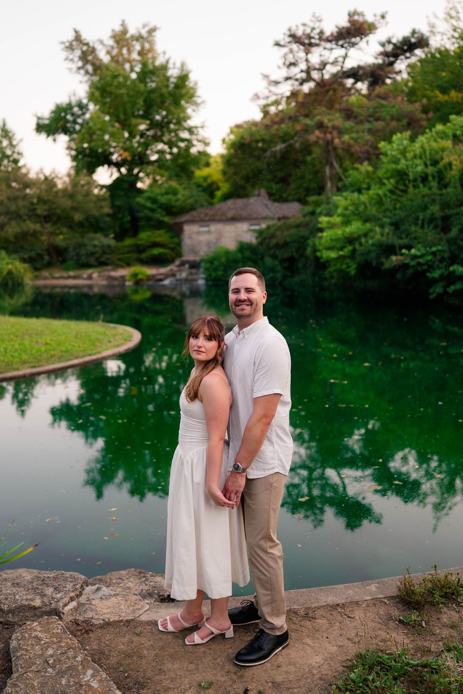 Couple holding hands standing on rocks beside a vibrant green reflective pond in a park.