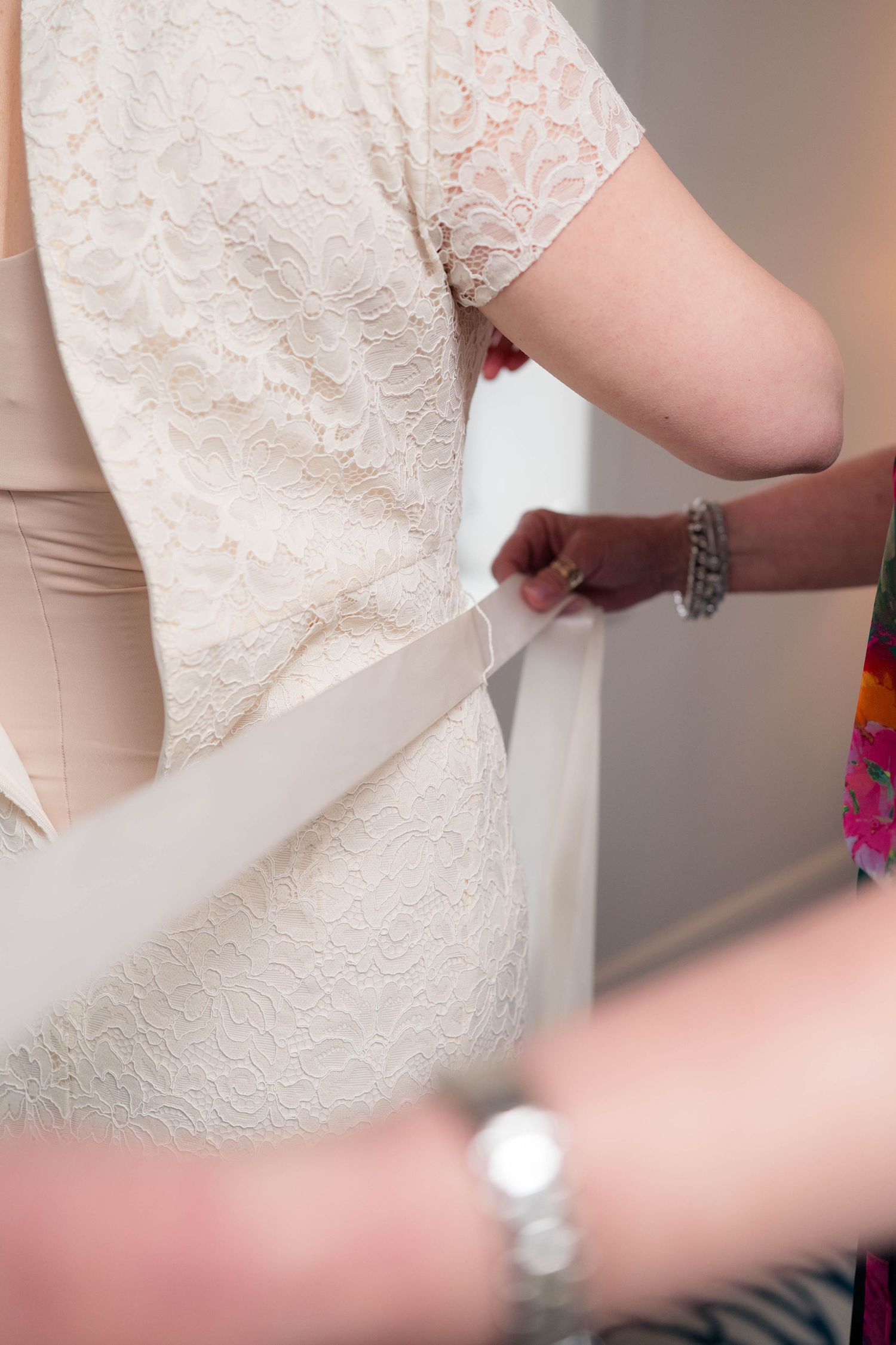 Helper ties a white satin sash around a bride in an ivory lace wedding dress.
