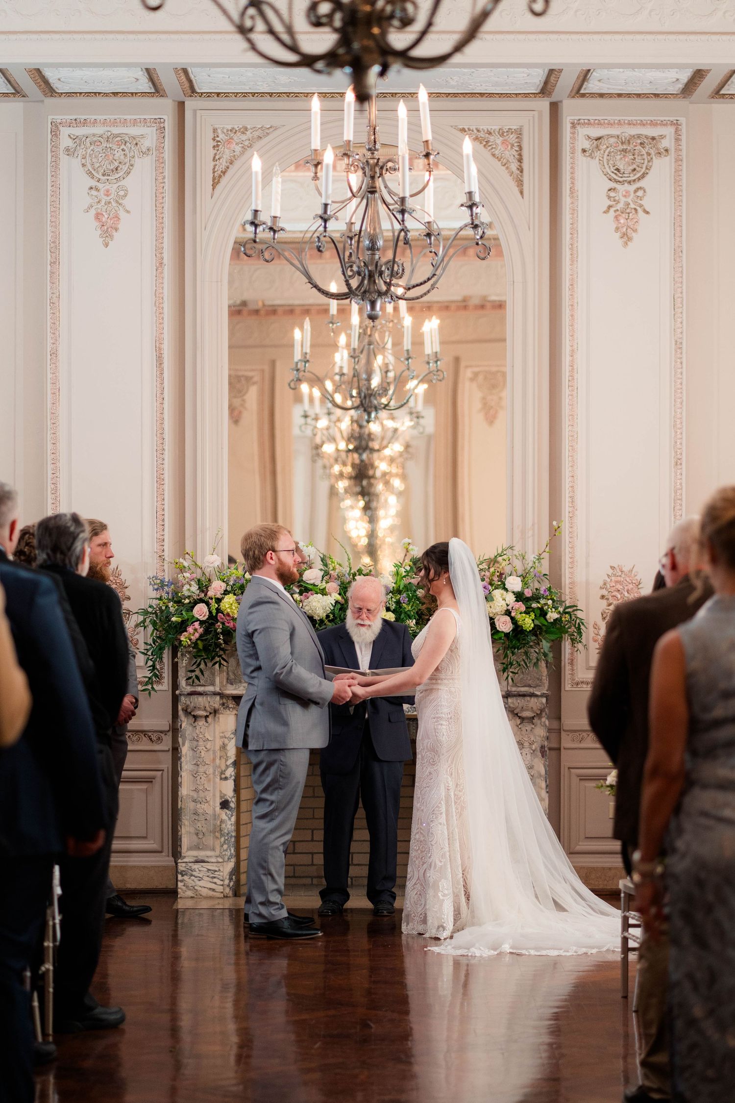 Couple exchanging vows at altar with floral arrangements under a chandelier in a ballroom.