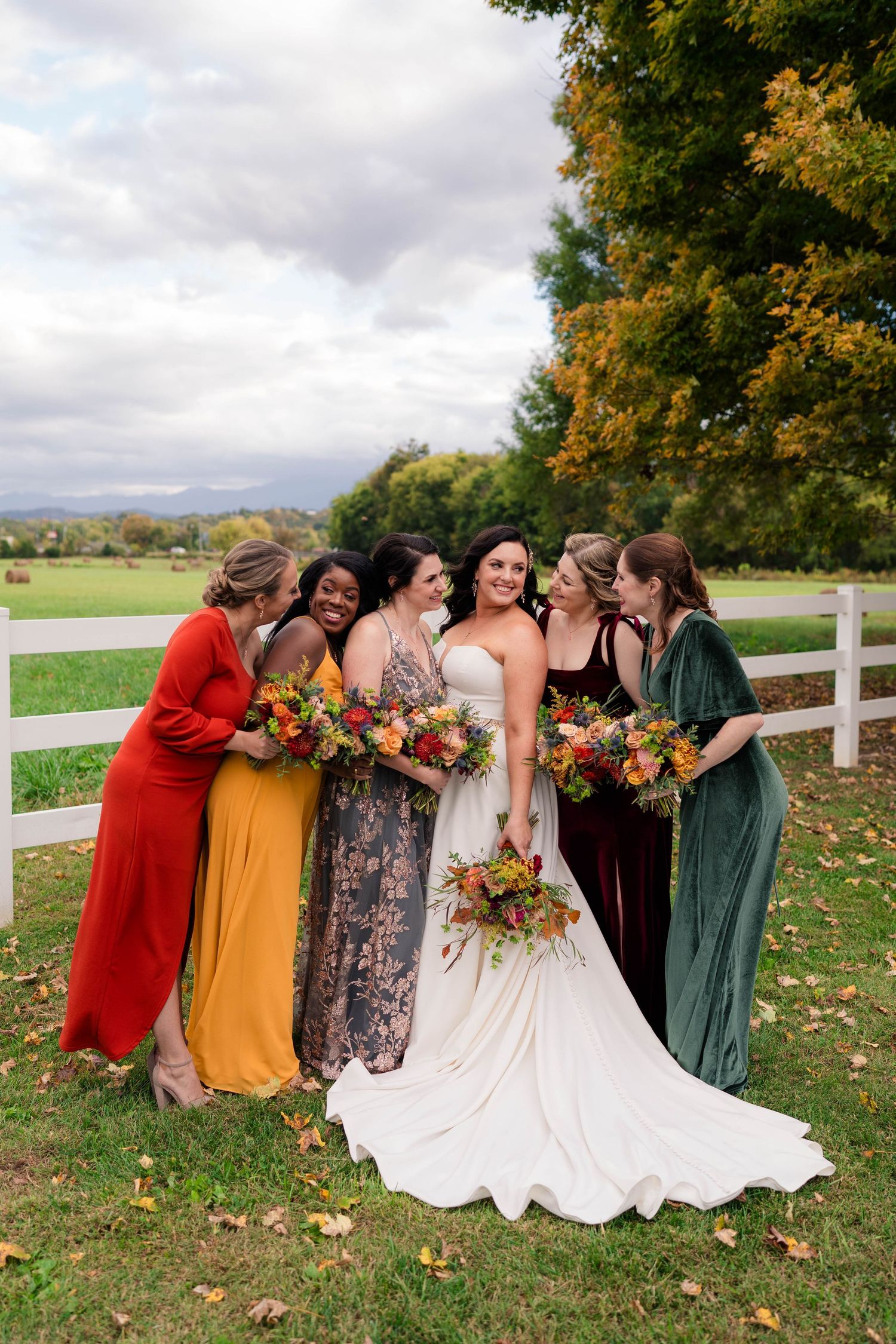 Bride surrounded by bridesmaids in mismatched fall-colored gowns holding vibrant autumn bouquets outdoors.
