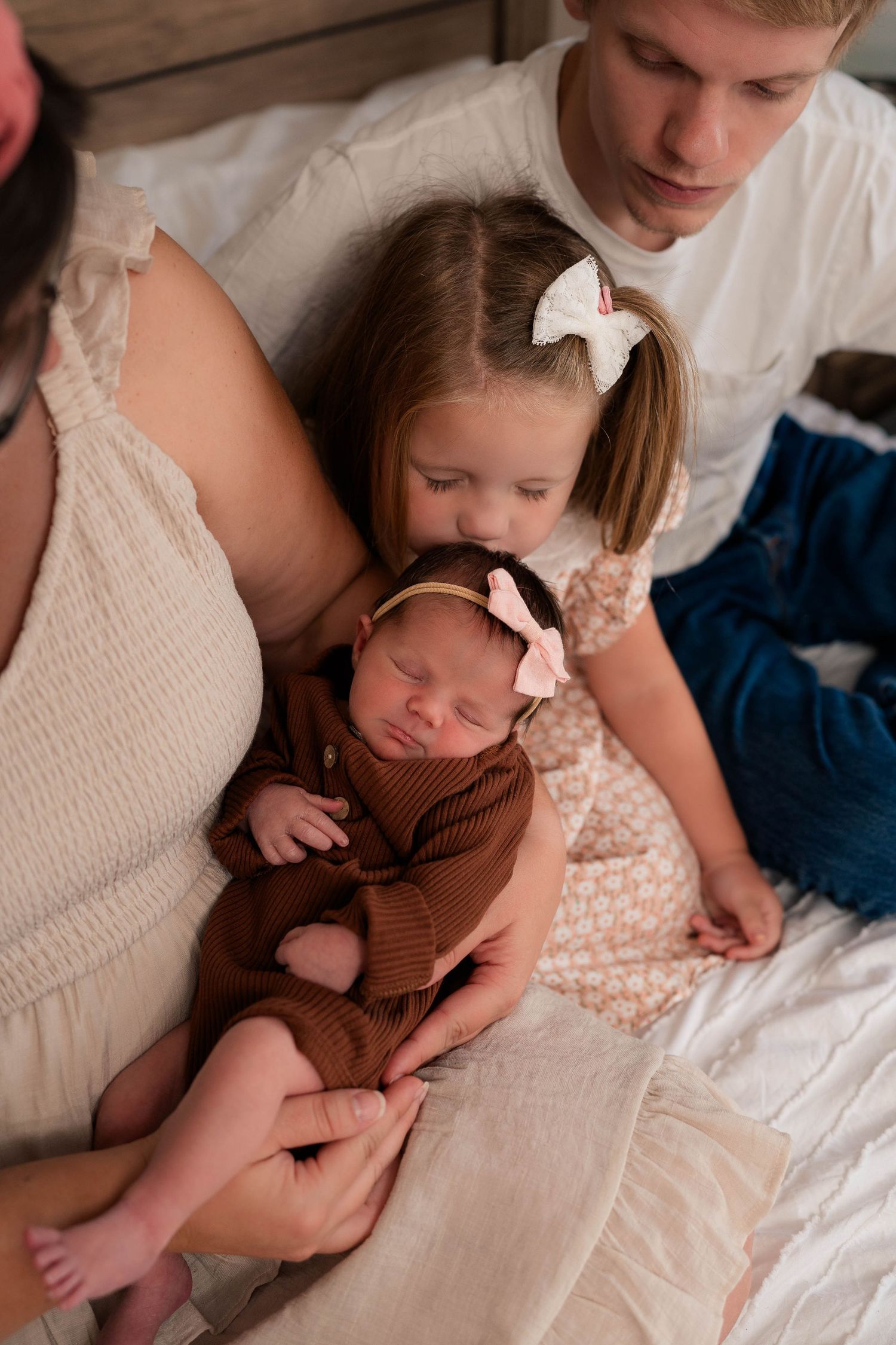 Family with newborn baby in brown outfit, toddler girl with bow, and older boy sitting together on a bed.
