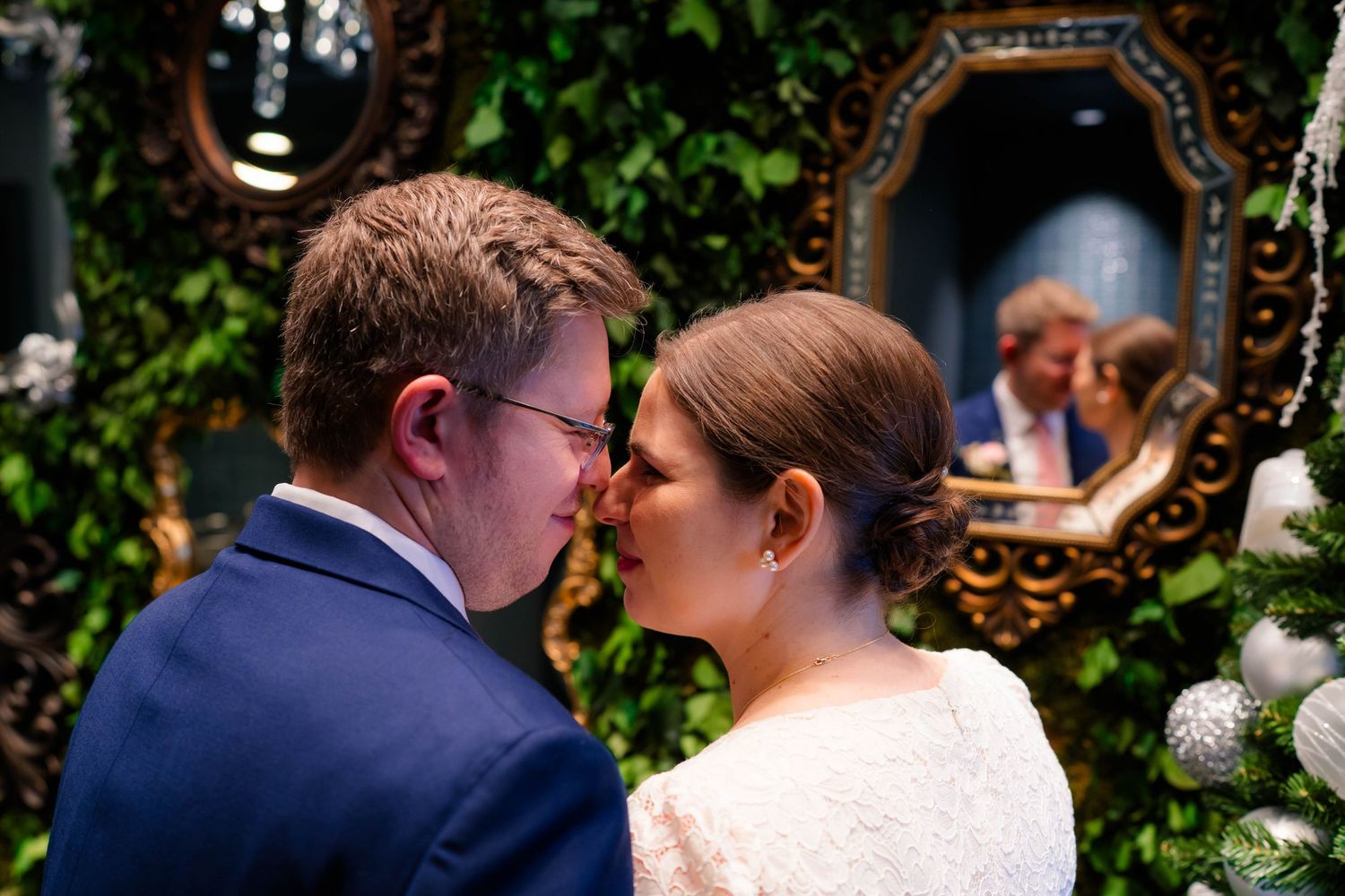 Bride and groom share a tender kiss in front of a green ivy wall with an ornate vintage mirror.