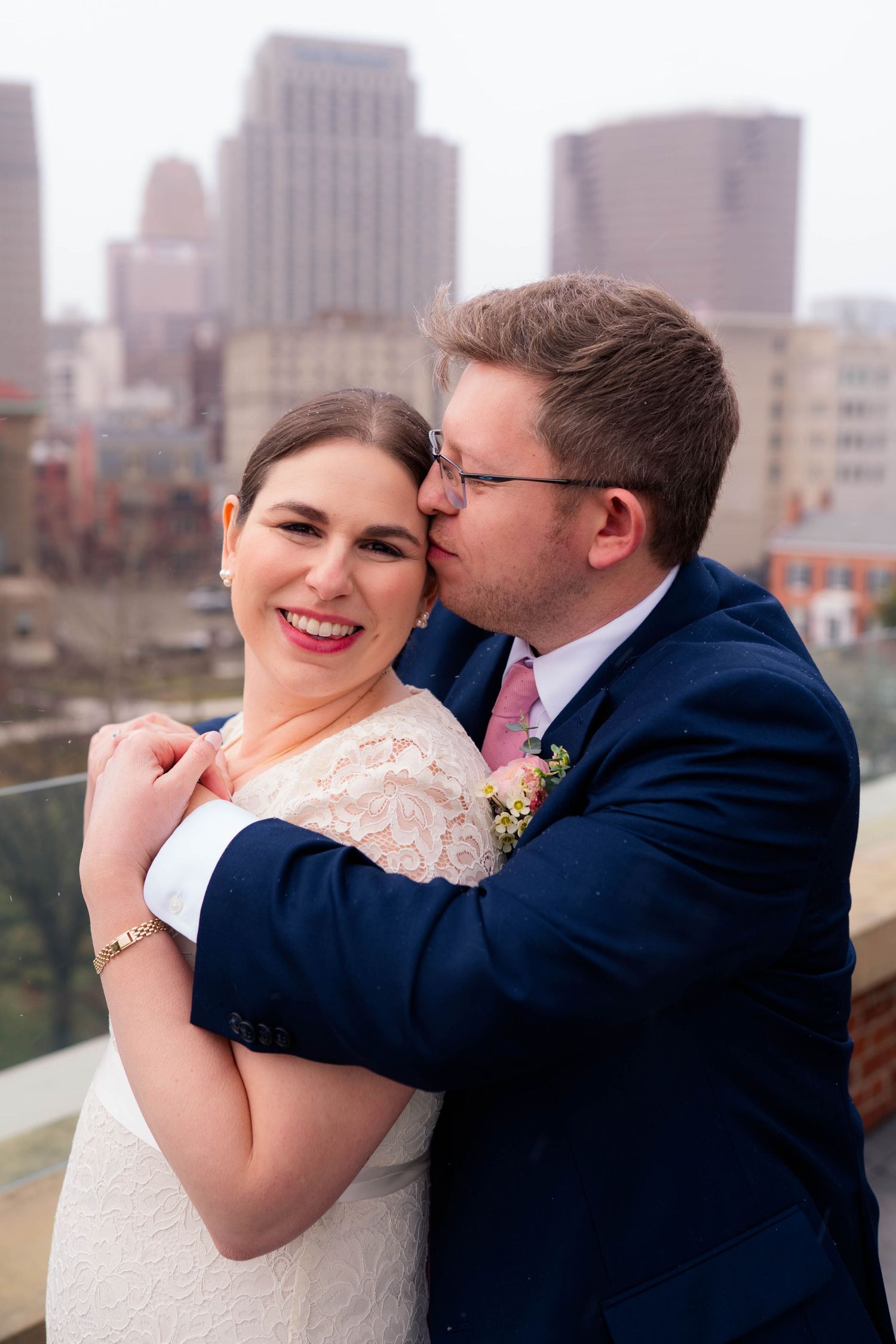 Groom in navy suit kissing bride on cheek as she smiles with city skyline in the background.