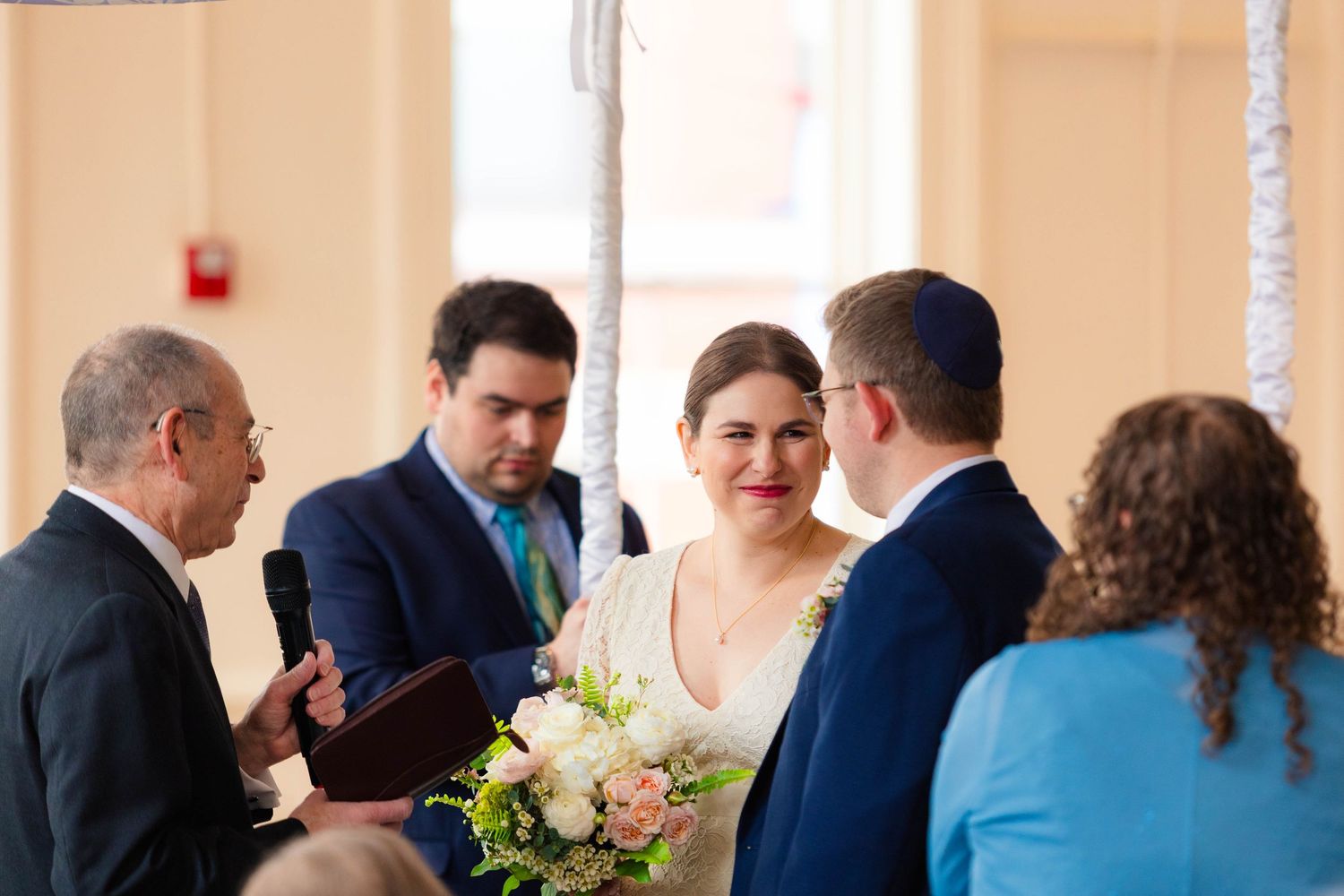 Smiling bride holding bouquet stands with groom and officiant during indoor wedding ceremony.