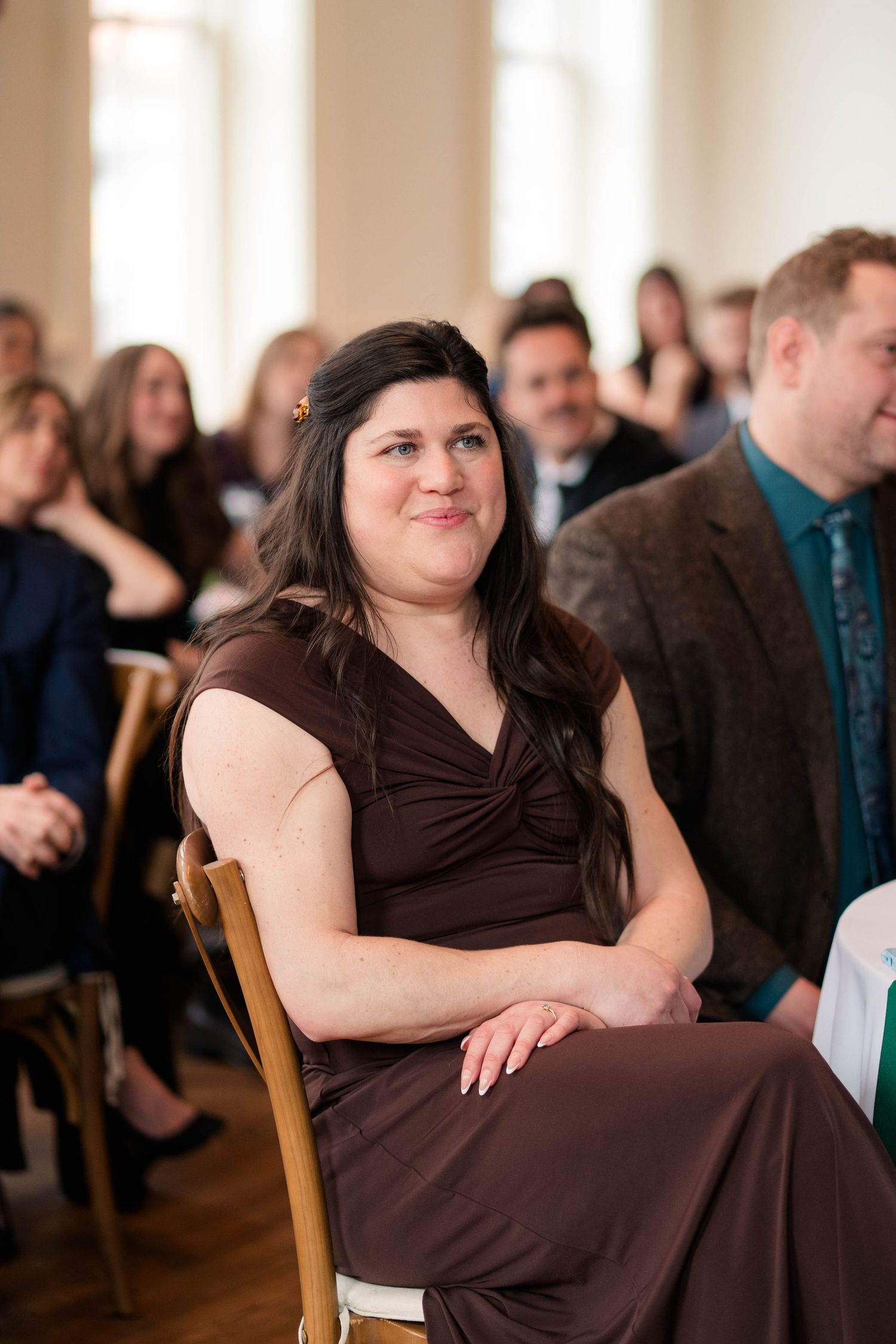 Dark-haired woman in brown dress smiles while seated among guests at wedding ceremony.