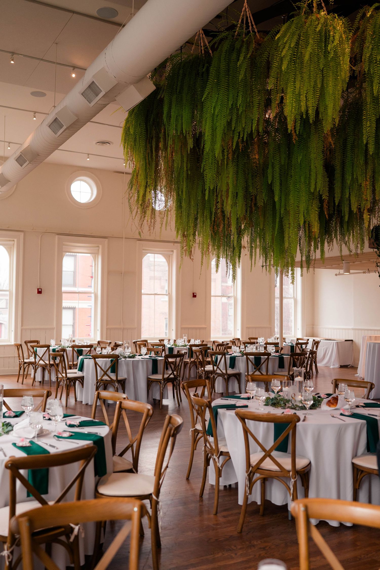 Elegant wedding reception hall with round tables, emerald green napkins, and lush hanging fern installation overhead.
