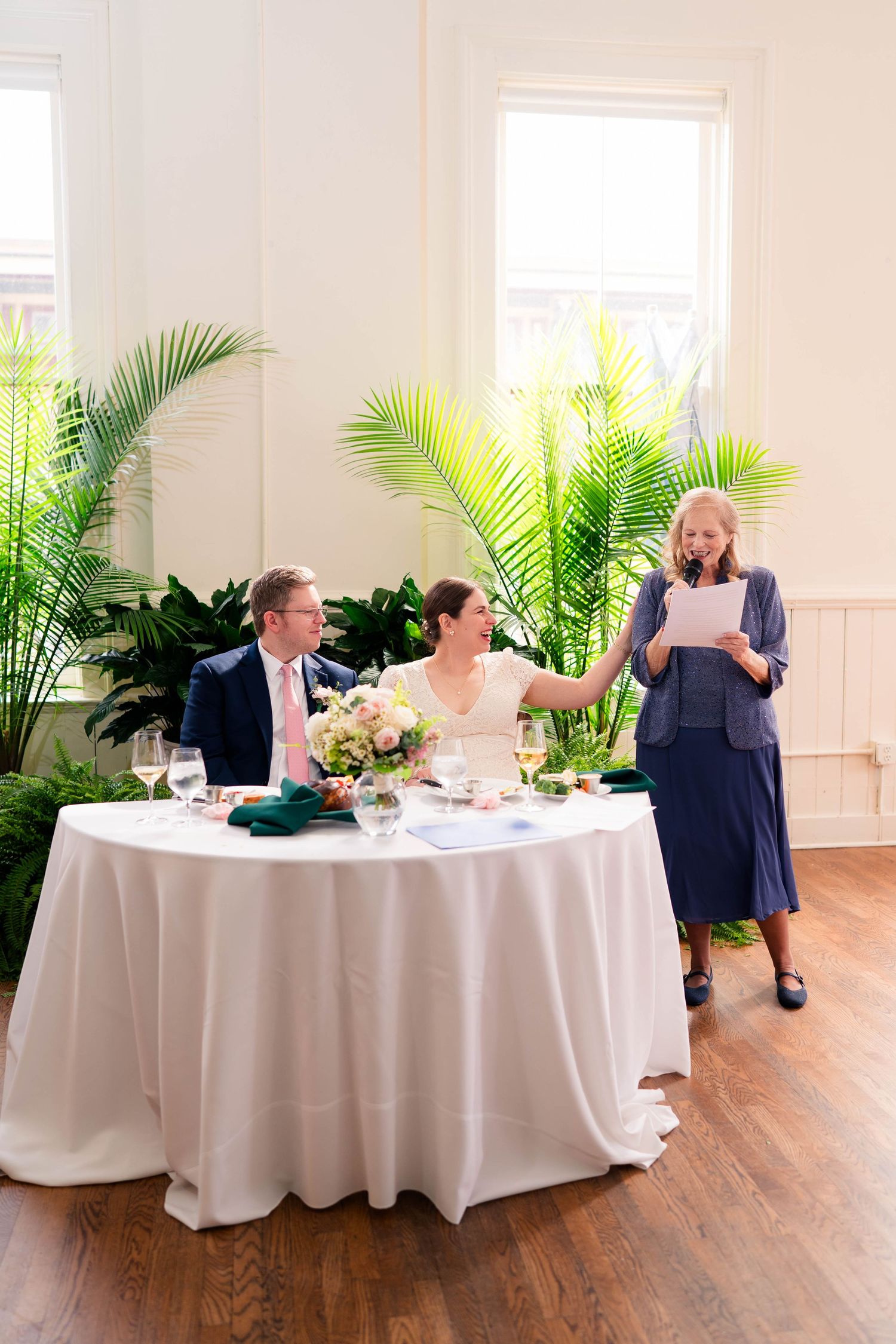 Woman in blue reads toast beside smiling bride and groom at sweetheart table.