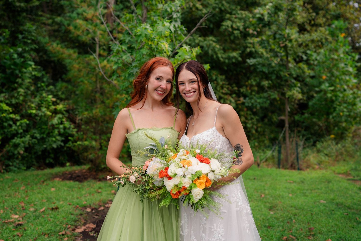 Bride holding a colorful bouquet poses with a bridesmaid in a sage green dress outdoors.