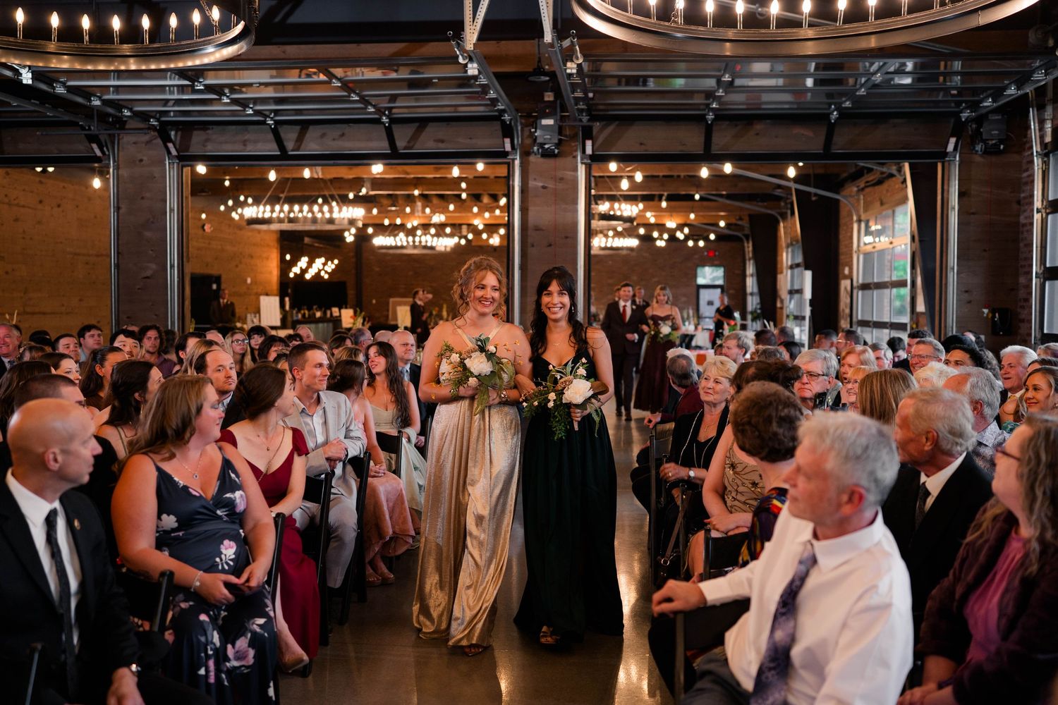 Bridesmaids in gold and black gowns walking down the aisle in an industrial wedding venue.