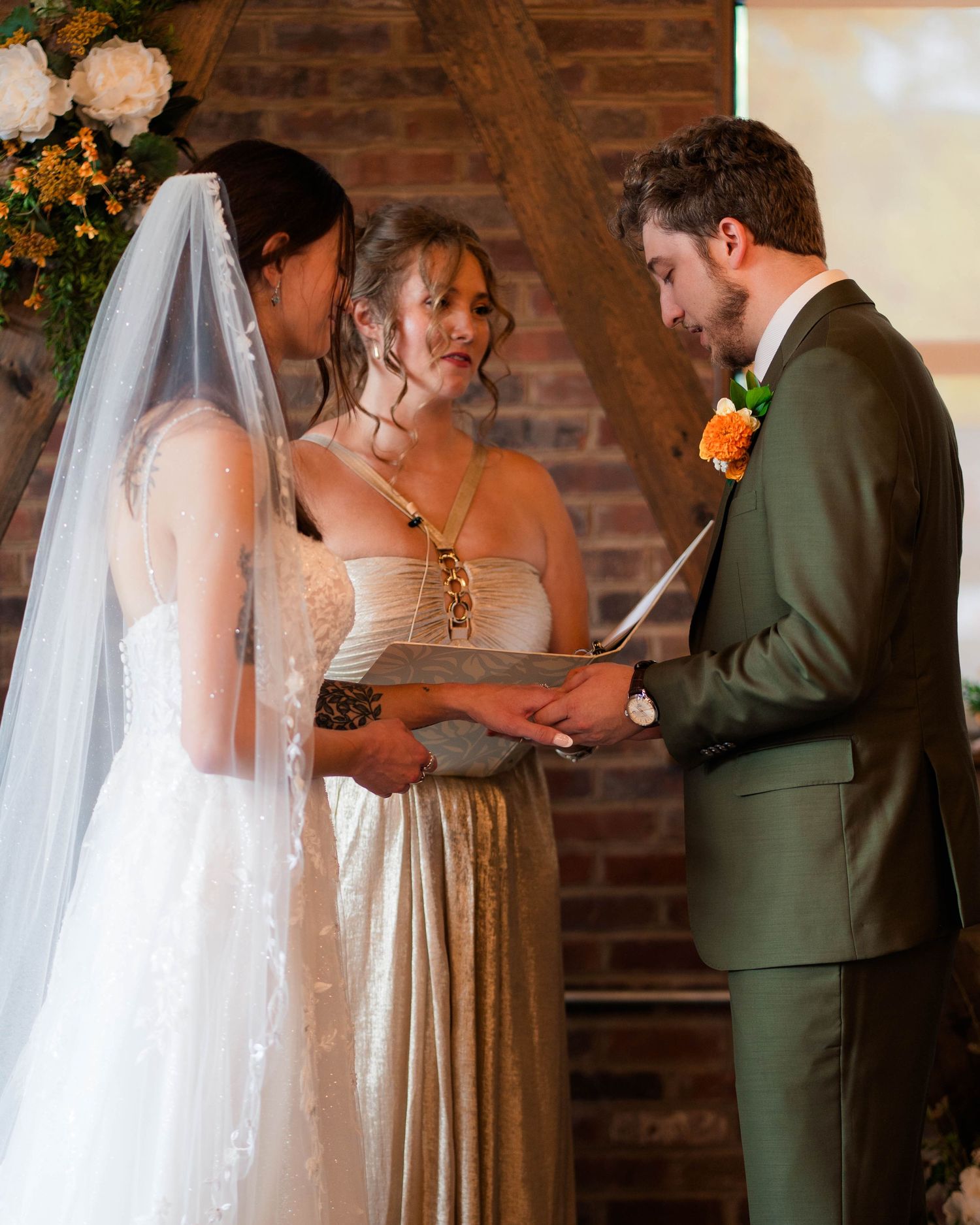 Groom in olive suit reads vows to bride in white gown during indoor wedding ceremony.