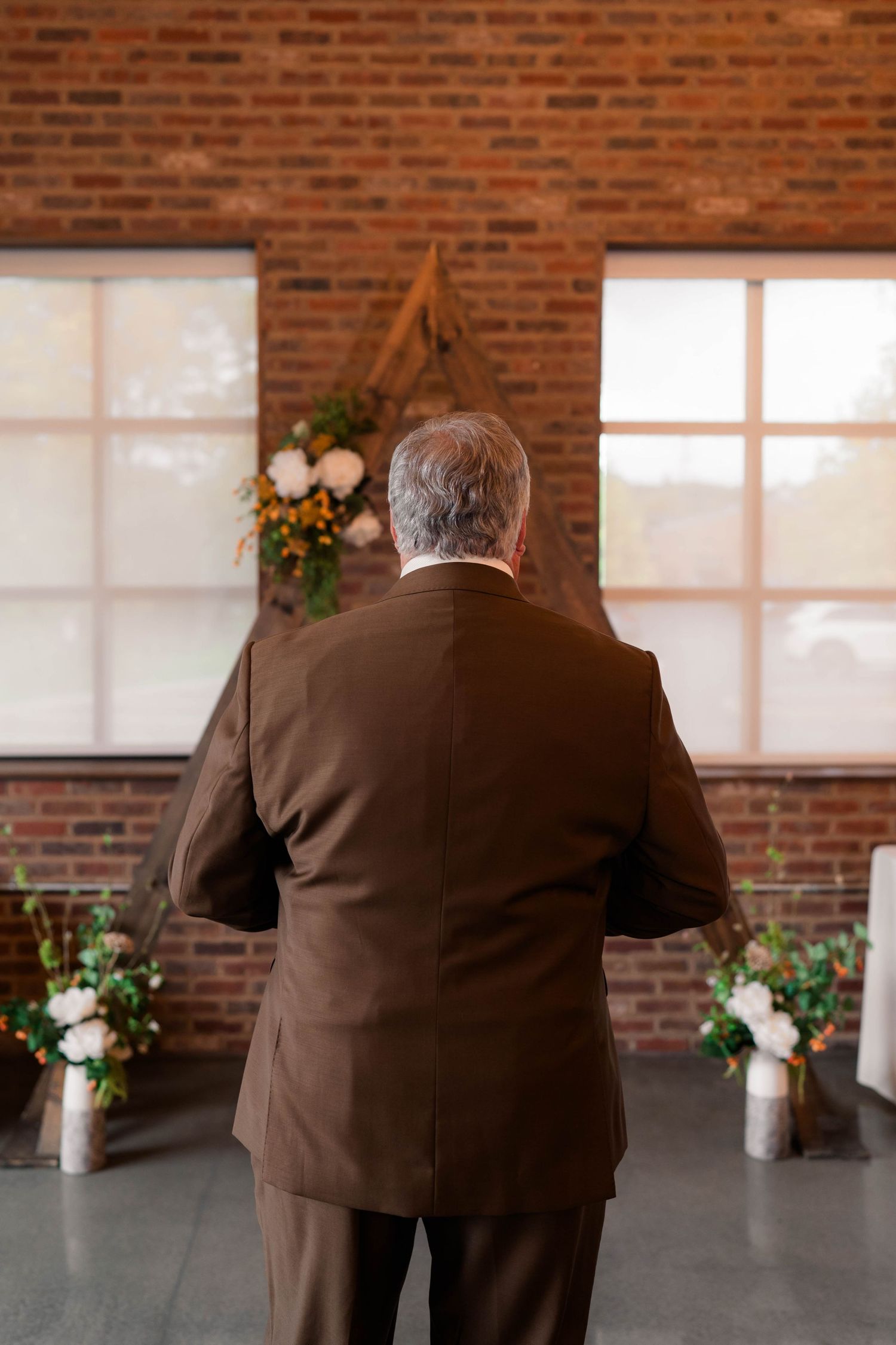 Man in brown suit stands at wedding altar with brick wall, floral arrangements, and triangular wooden arch.