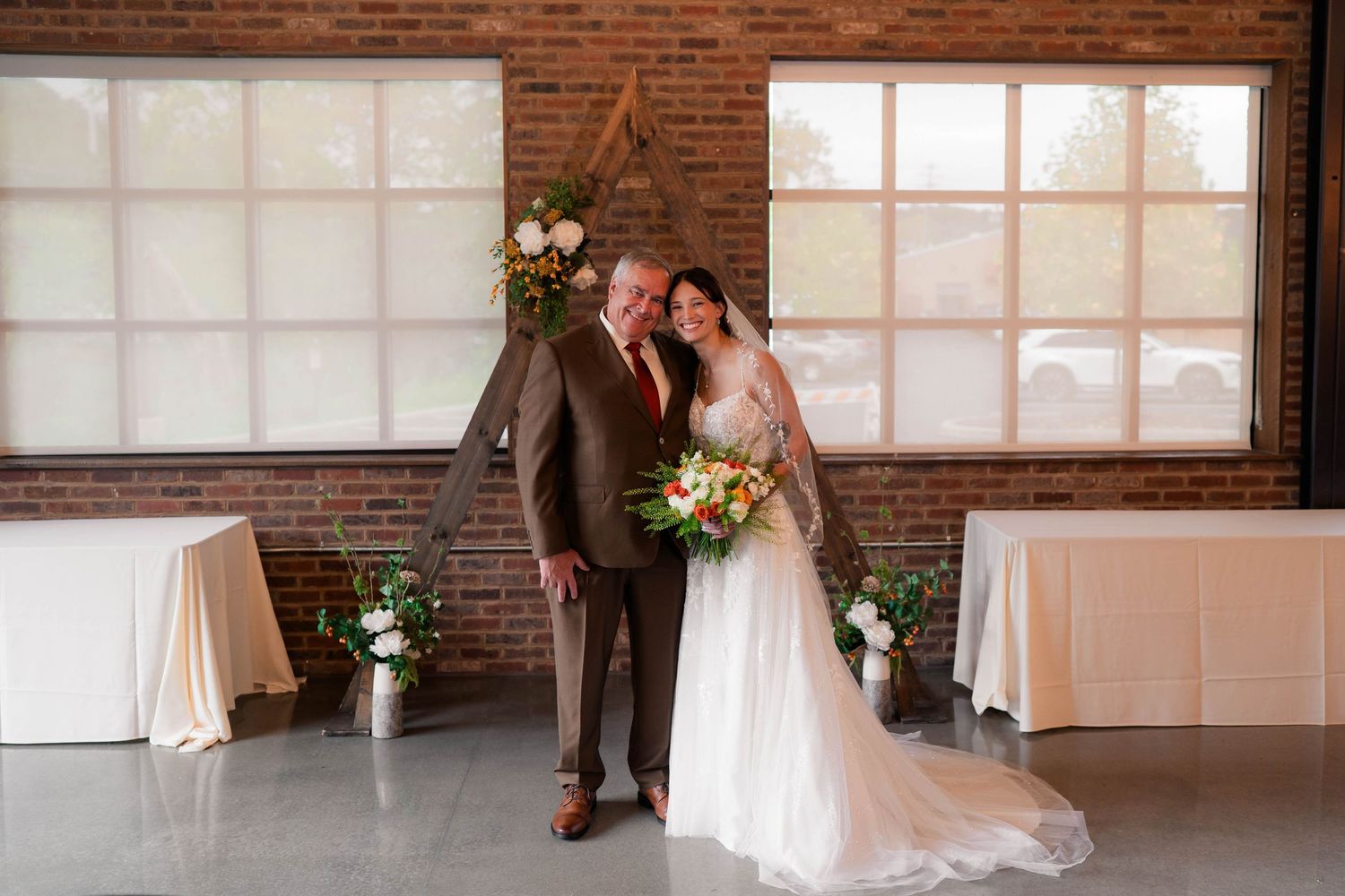 A bride holding a floral bouquet poses with an older man in a brick-walled venue.