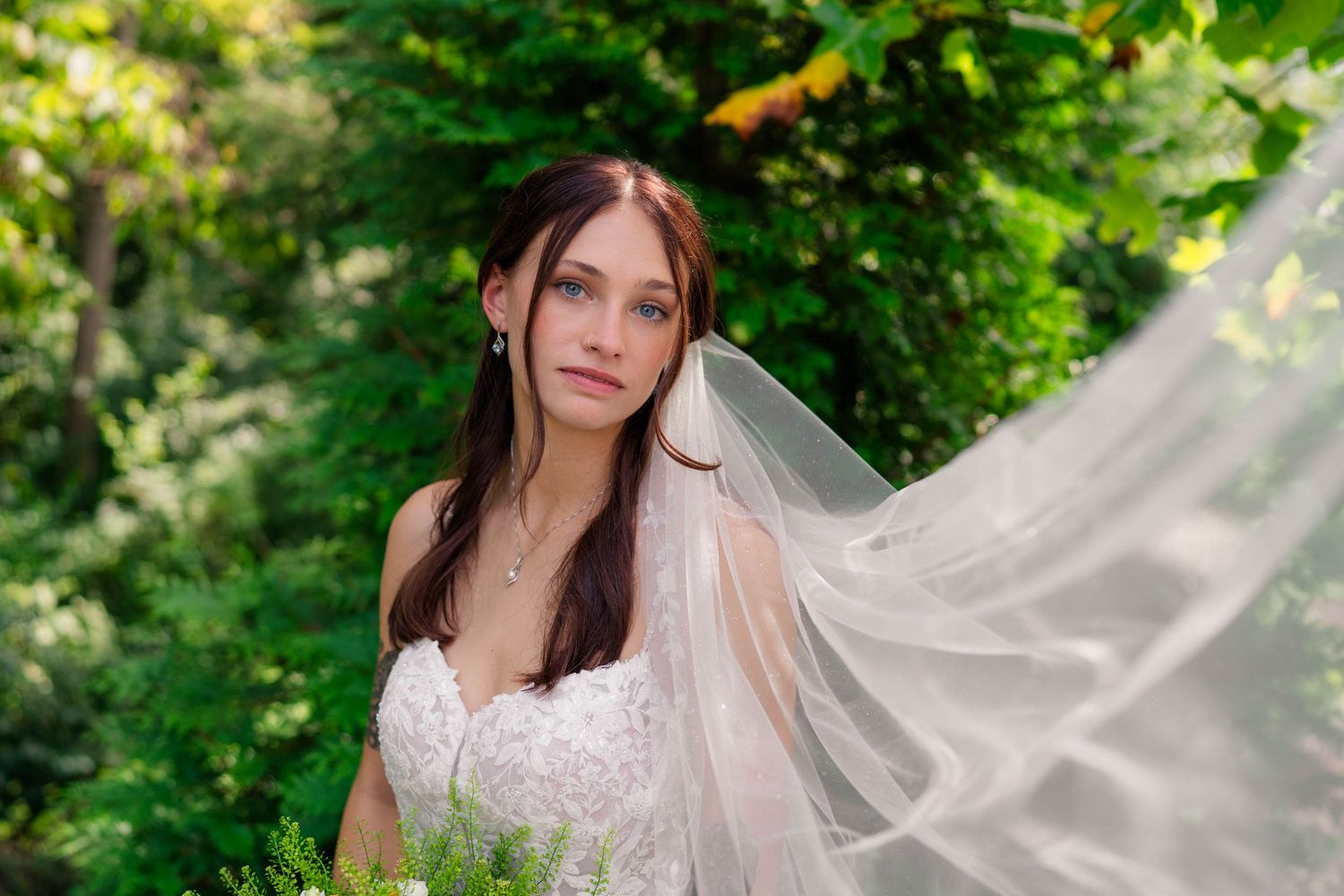 Beautiful bride in white gown with flowing veil surrounded by lush green foliage.