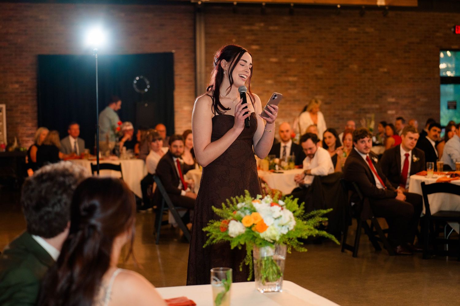 Woman in black dress giving maid of honor speech at wedding reception with seated guests.
