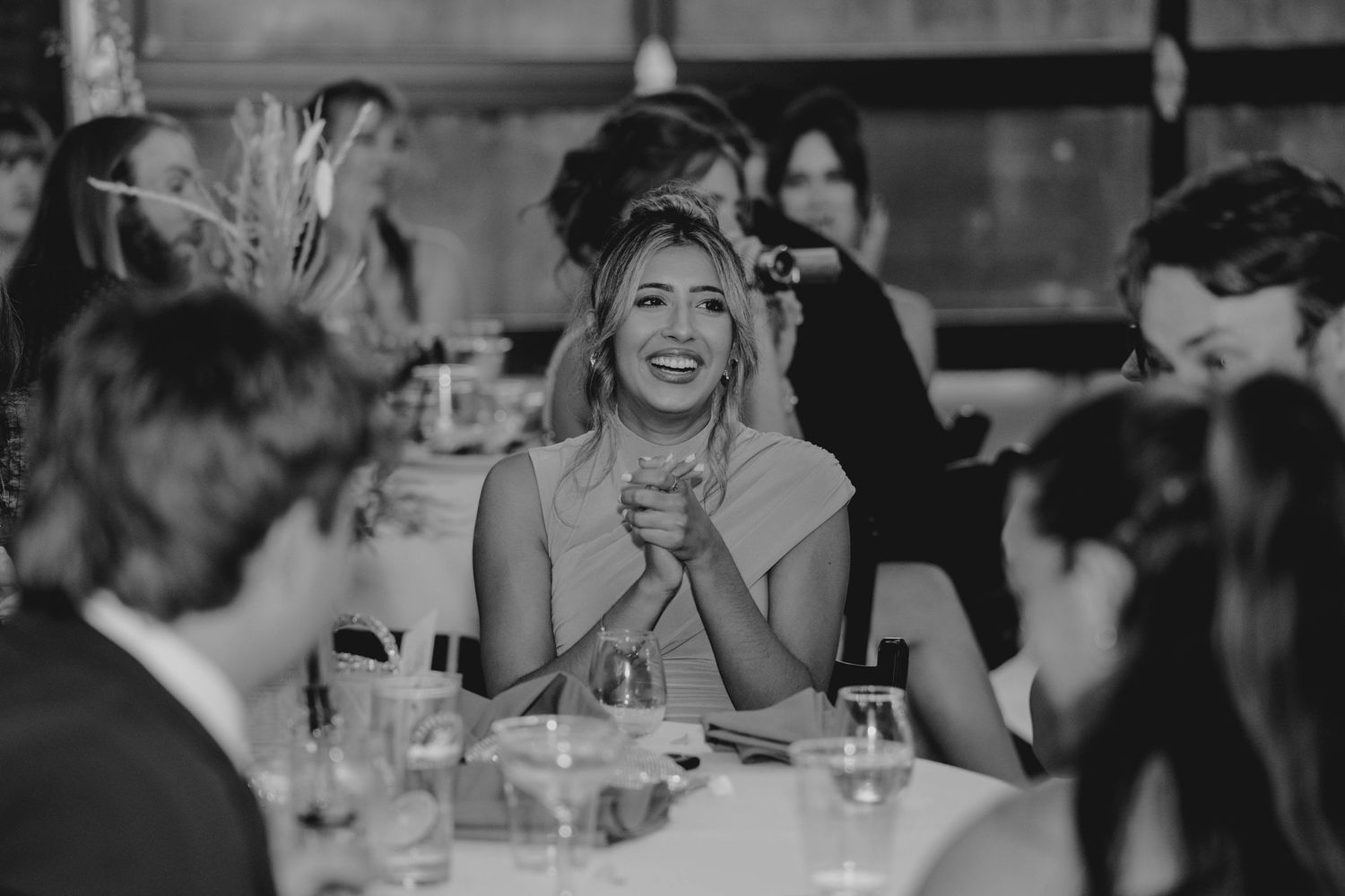 Black and white photo of joyful wedding guest laughing with clasped hands at reception dinner.