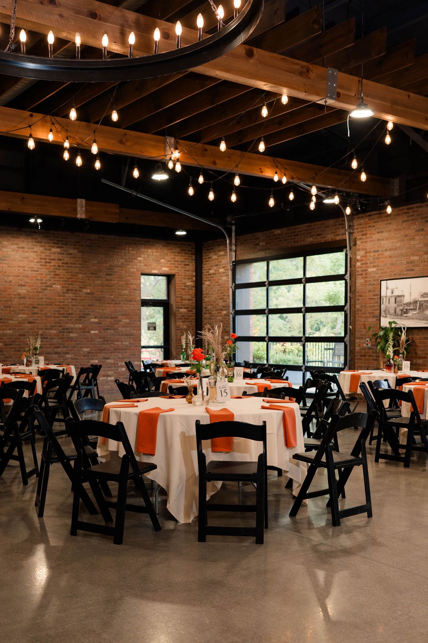 Industrial wedding venue with Edison bulb lighting, brick walls, black folding chairs, and tables with white linens and orange napkins.