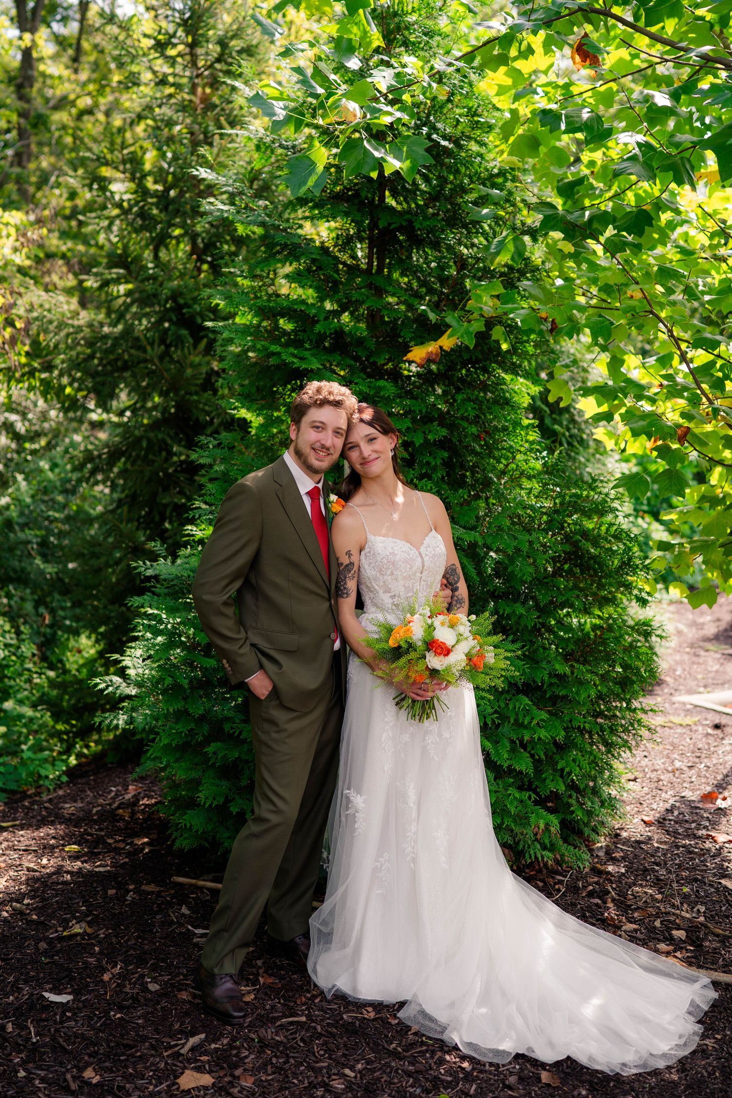 Bride and groom pose together in lush green garden, bride holding colorful bouquet in white lace gown.