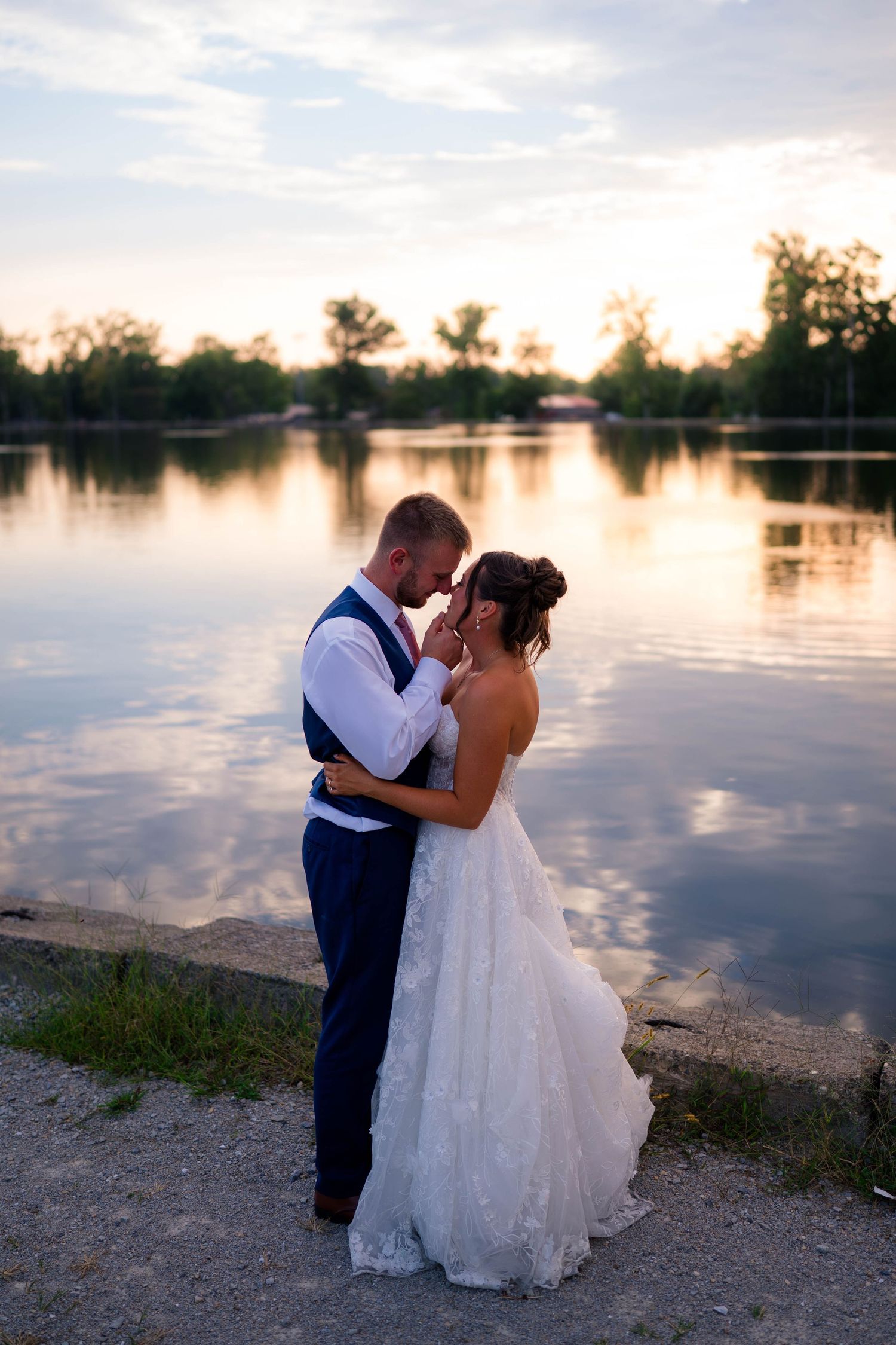 Bride and groom share a romantic embrace by a serene lake at sunset, with trees reflecting on calm water.