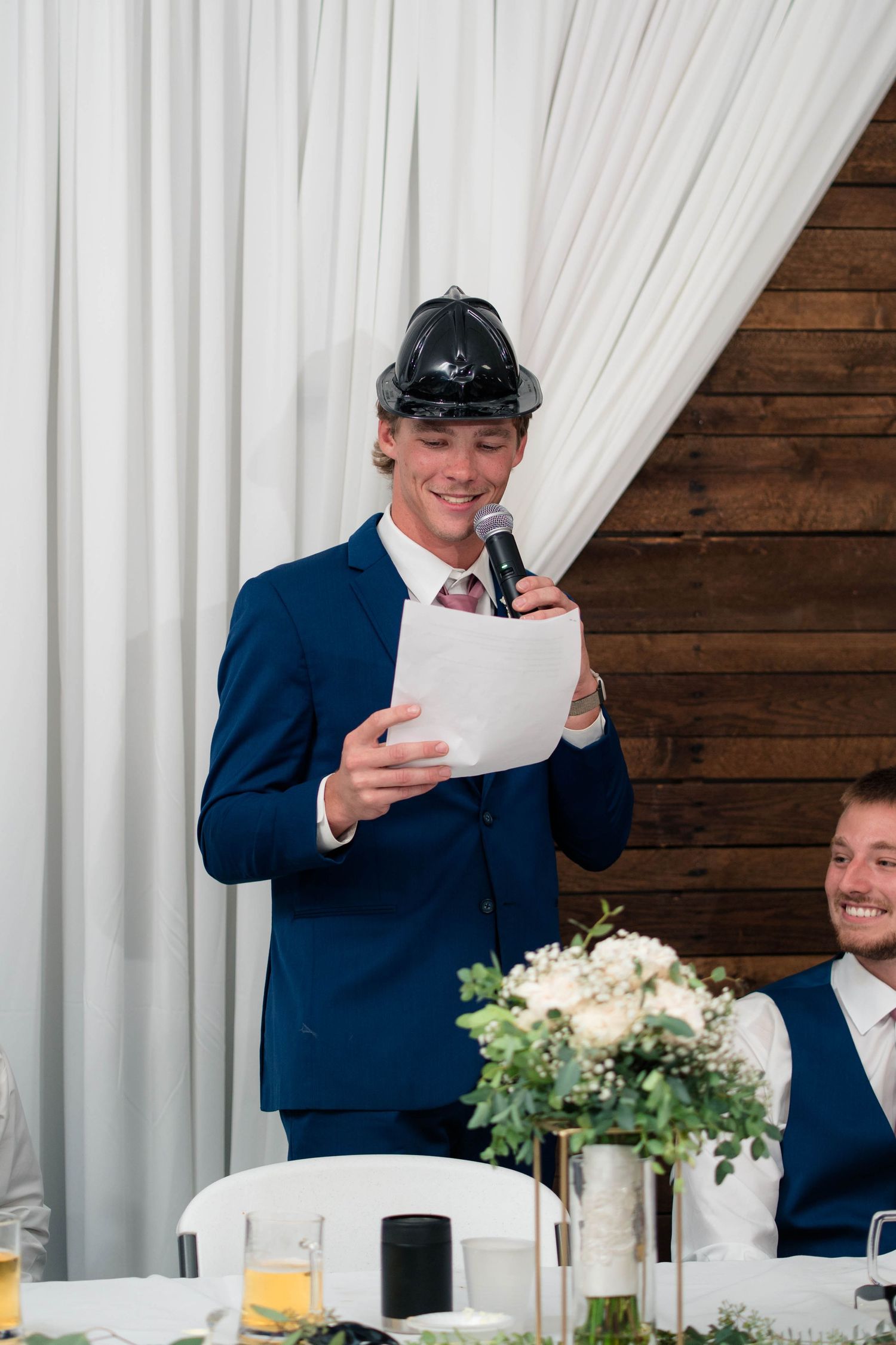 Best man in a hard hat reads a humorous wedding speech into a microphone at a reception.