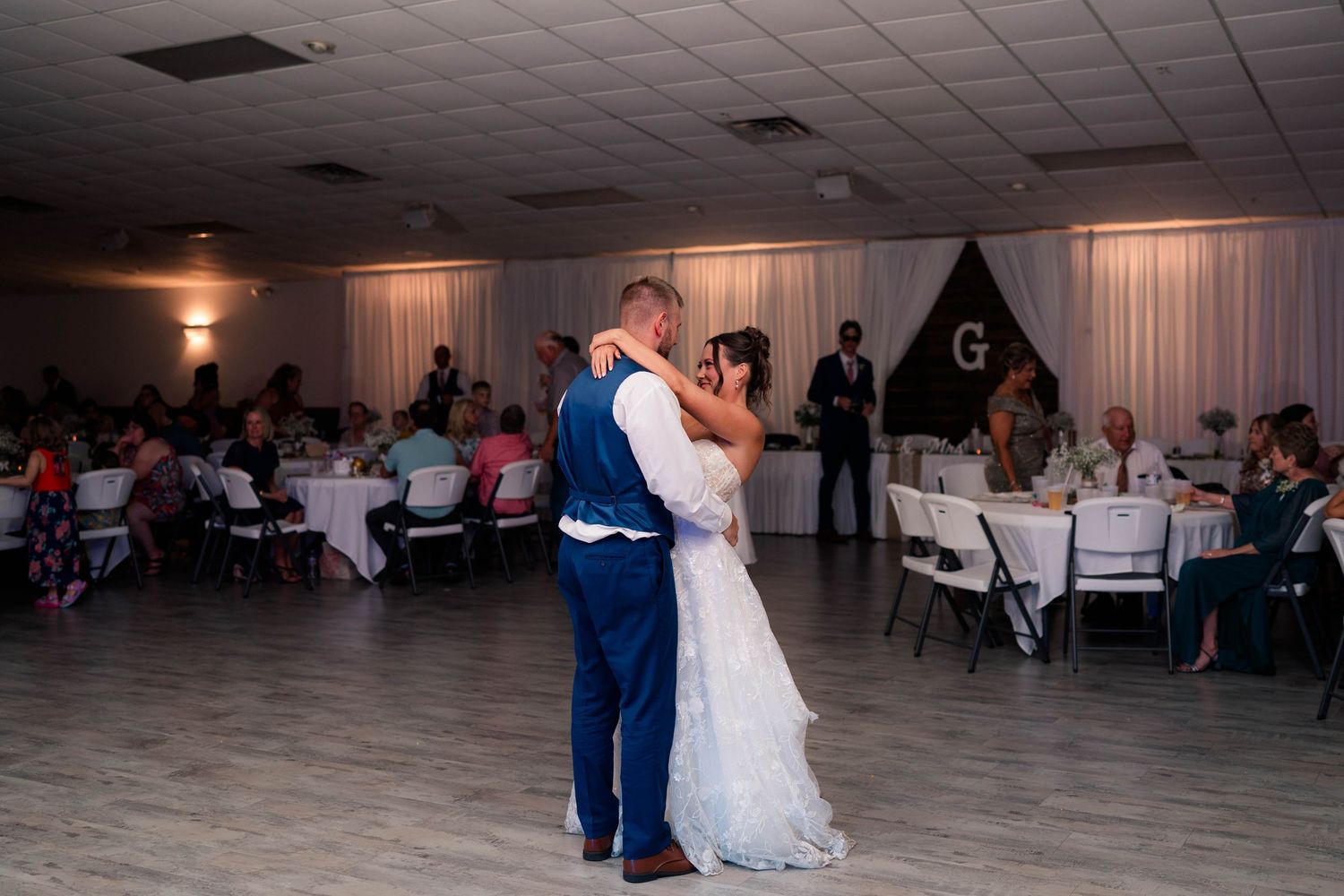 Bride and groom share romantic first dance on empty floor surrounded by seated wedding reception guests.