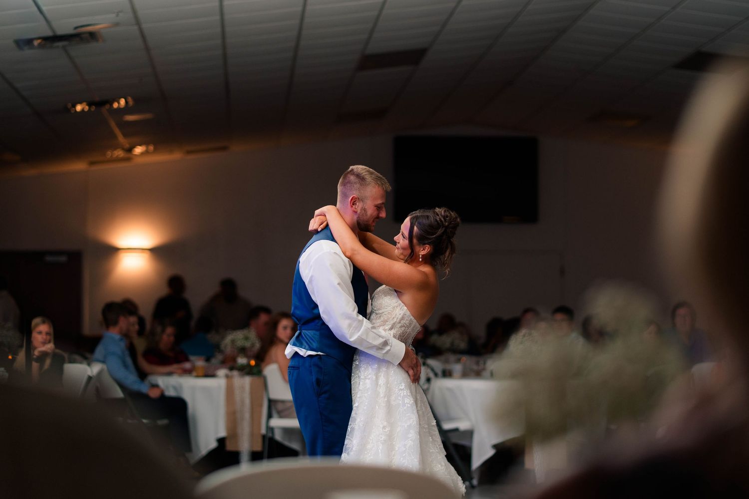 Bride and groom share an intimate first dance embrace in softly lit wedding reception hall.