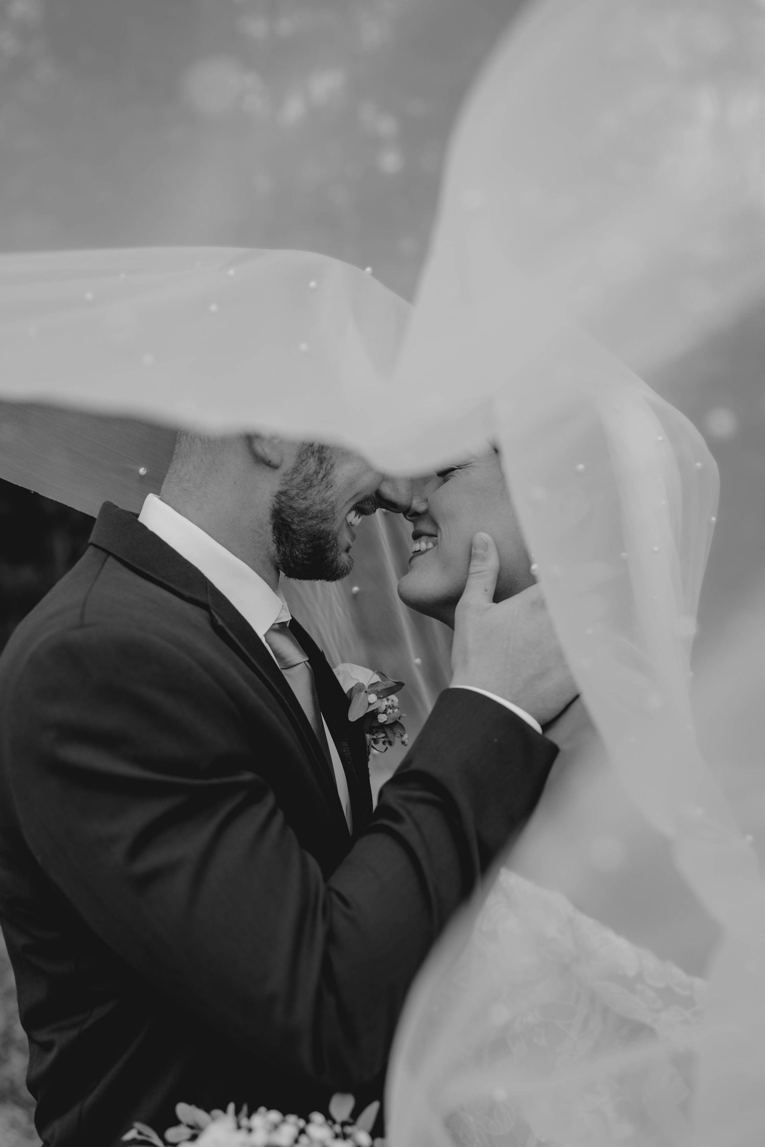 Romantic black and white photo of bride and groom almost kissing beneath a flowing wedding veil.