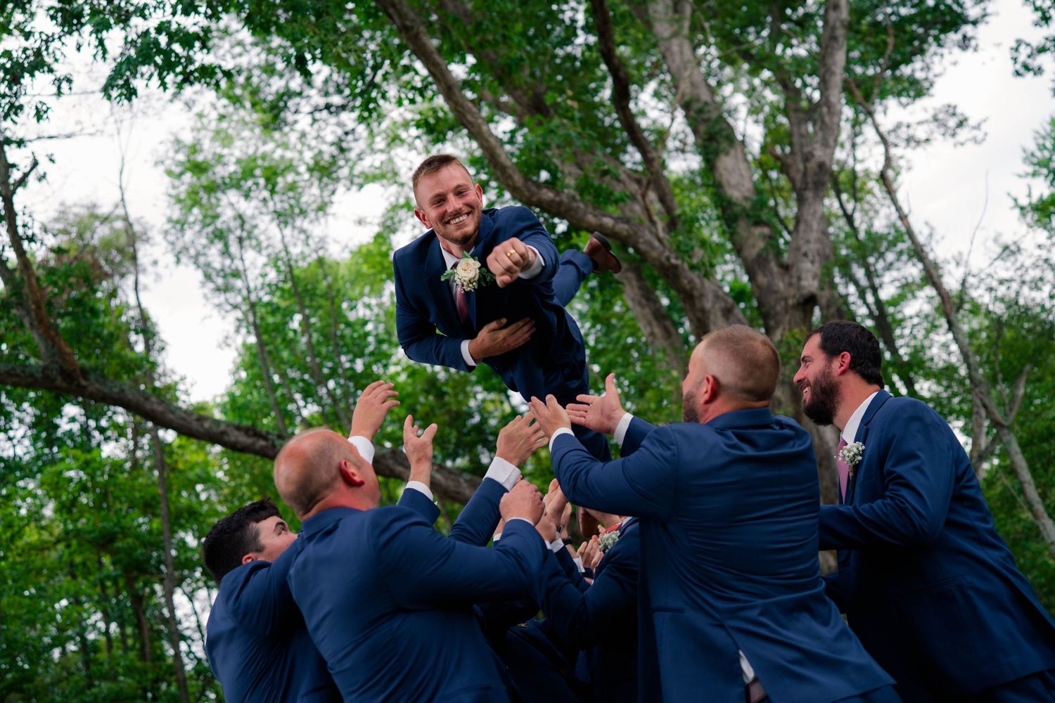 Groomsmen tossing smiling groom into the air in a celebratory moment outdoors among trees.