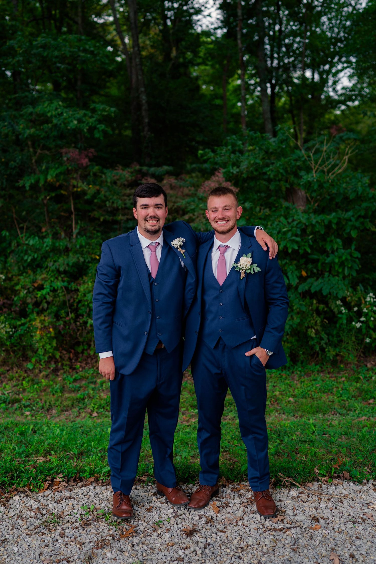 Two groomsmen in navy blue suits with pink ties and boutonnieres pose together outdoors at a wedding.