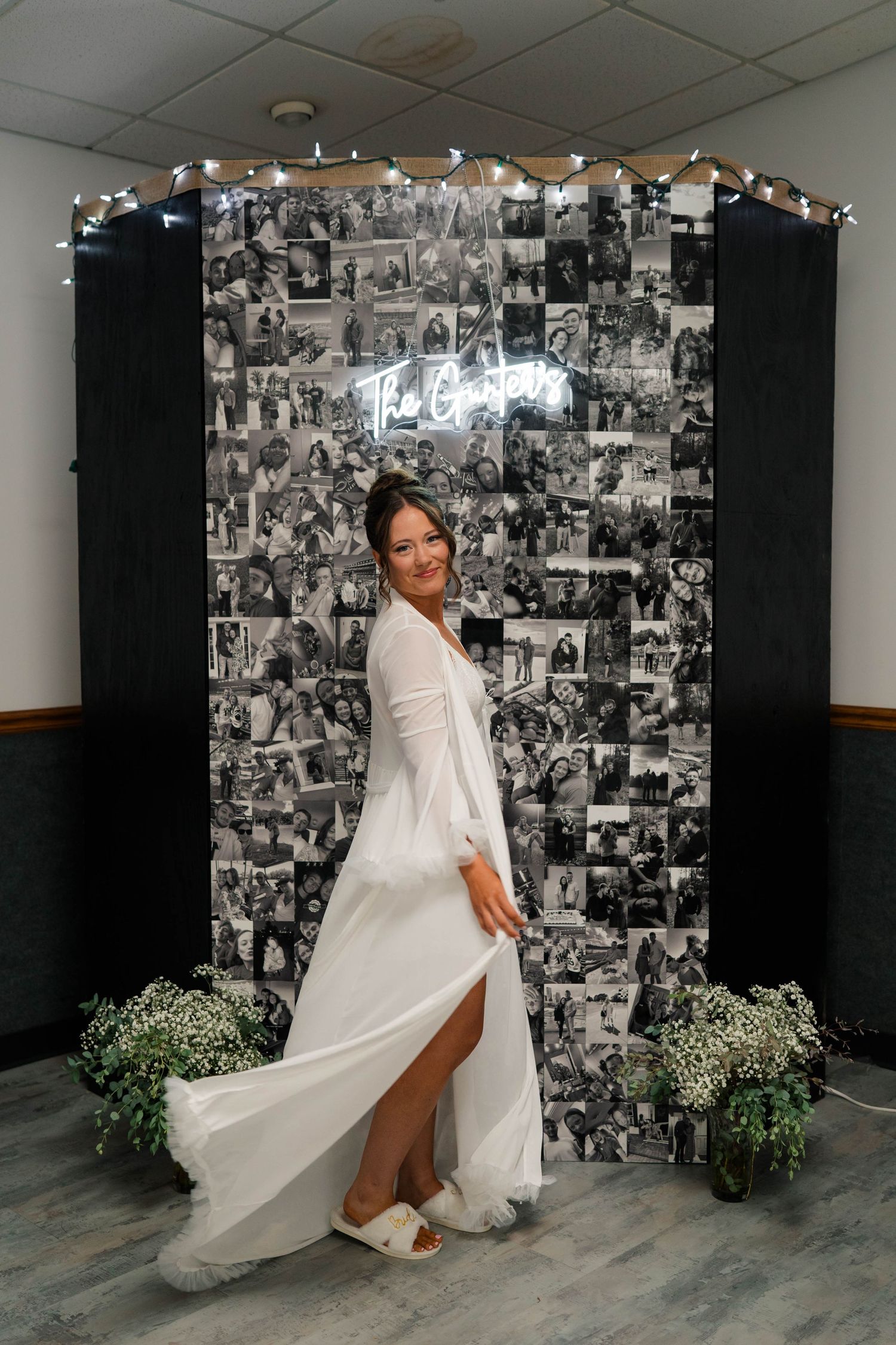 Bride in white gown twirling in front of black and white photo collage backdrop with neon sign and fairy lights.