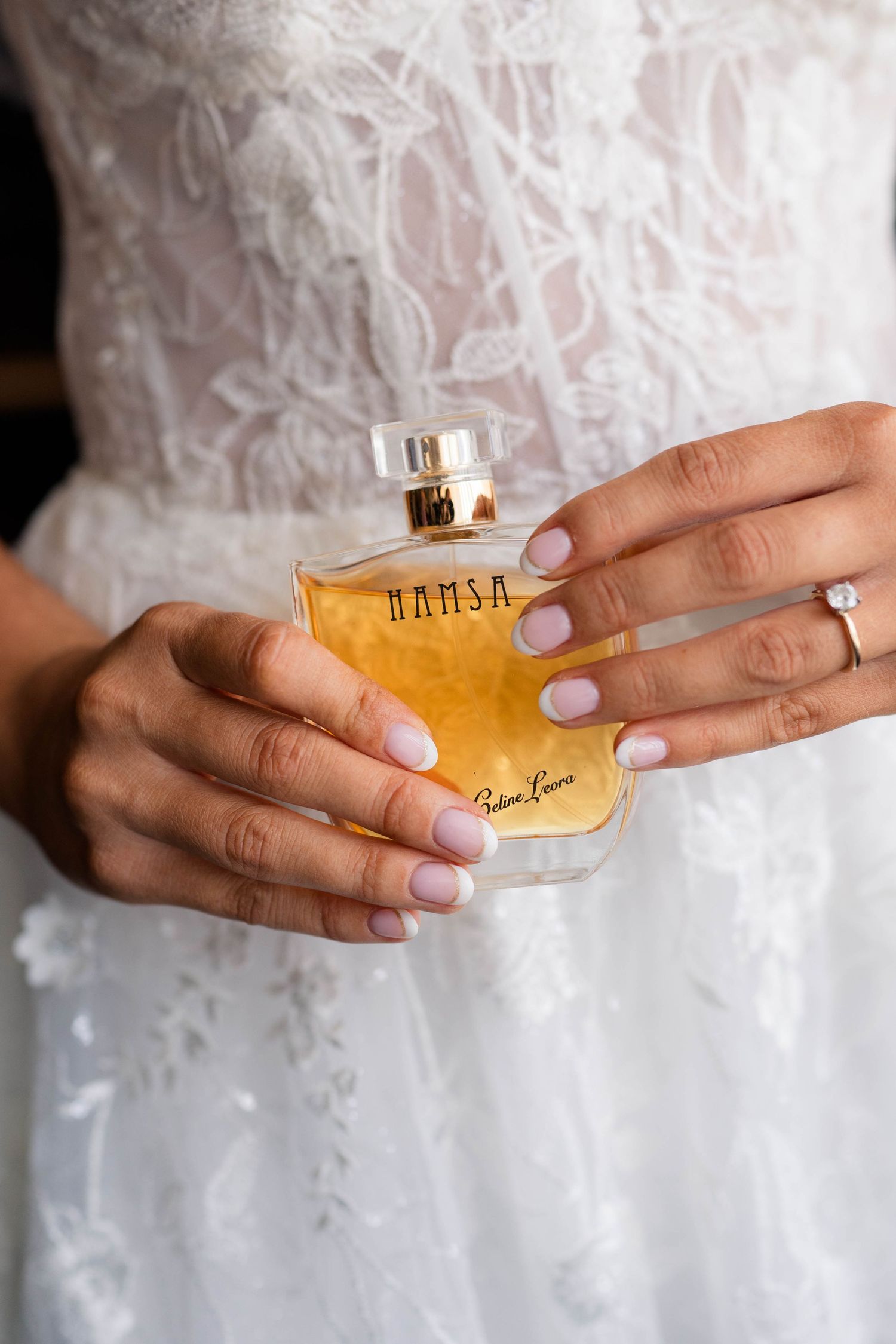 Bride holding Hamsa by Paloma Picasso perfume bottle wearing diamond engagement ring and lace gown.