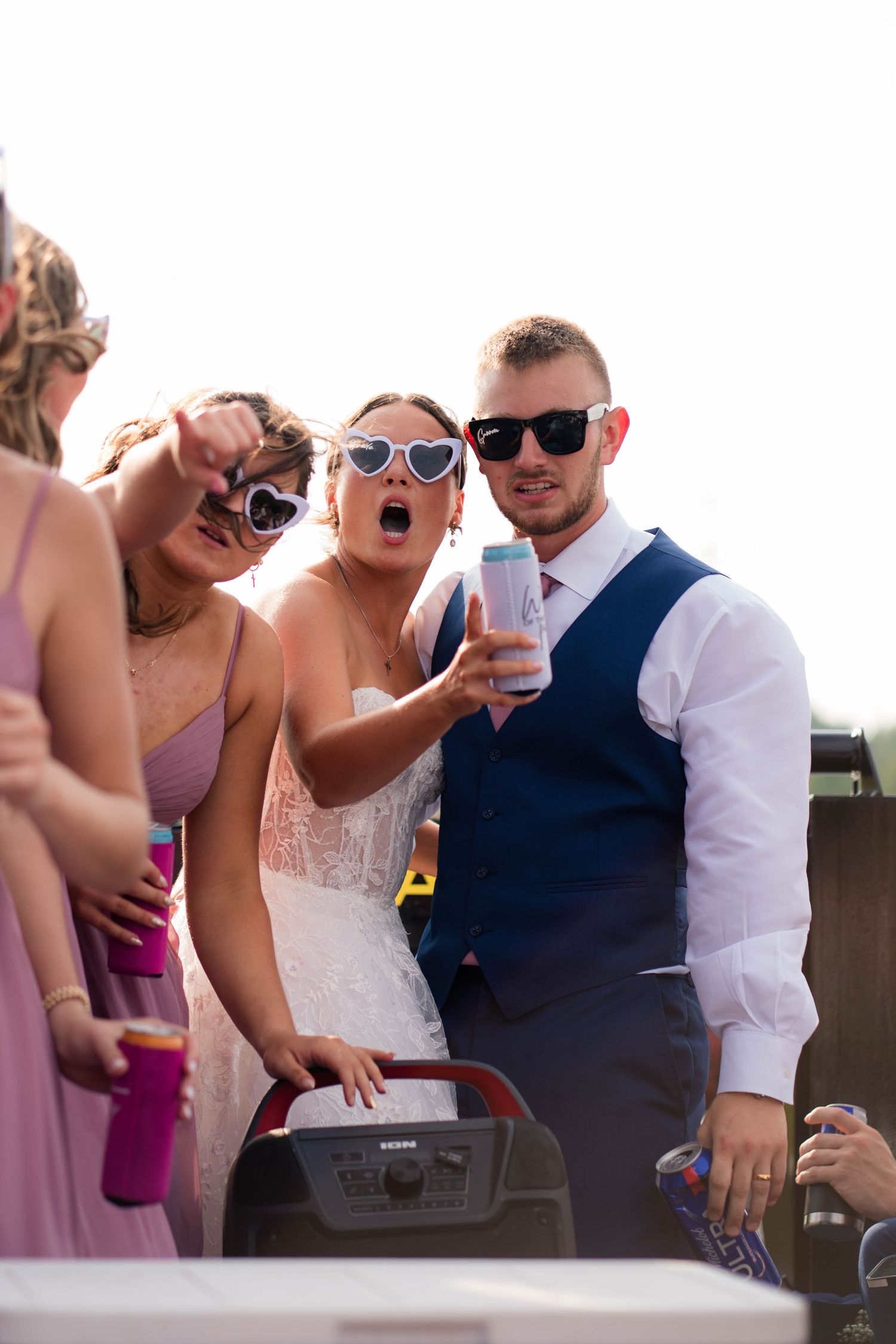 Bride in heart sunglasses and groom in navy vest celebrate with guests at outdoor wedding party.