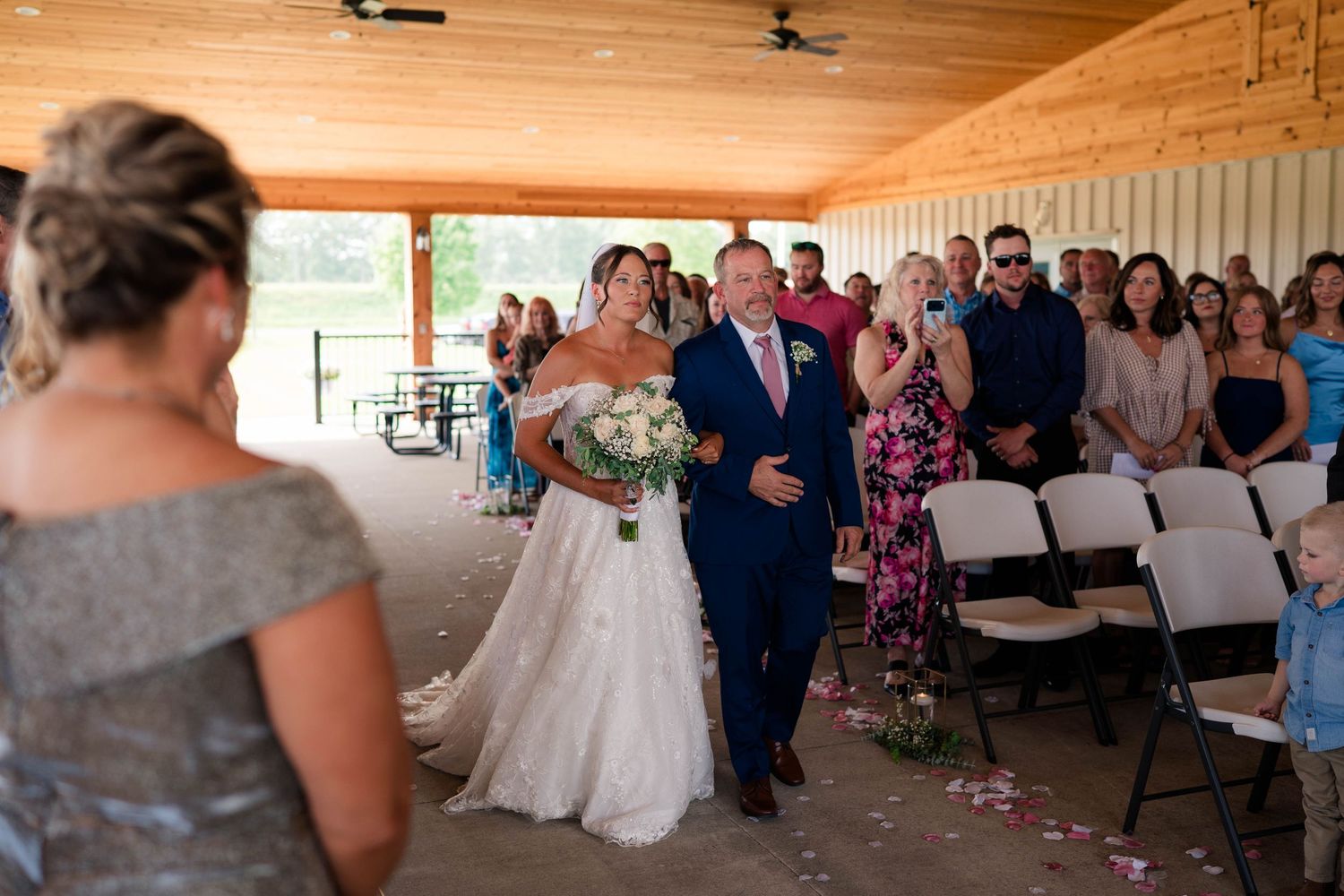Bride in lace gown walks down aisle with escort toward waiting groom at outdoor ceremony.