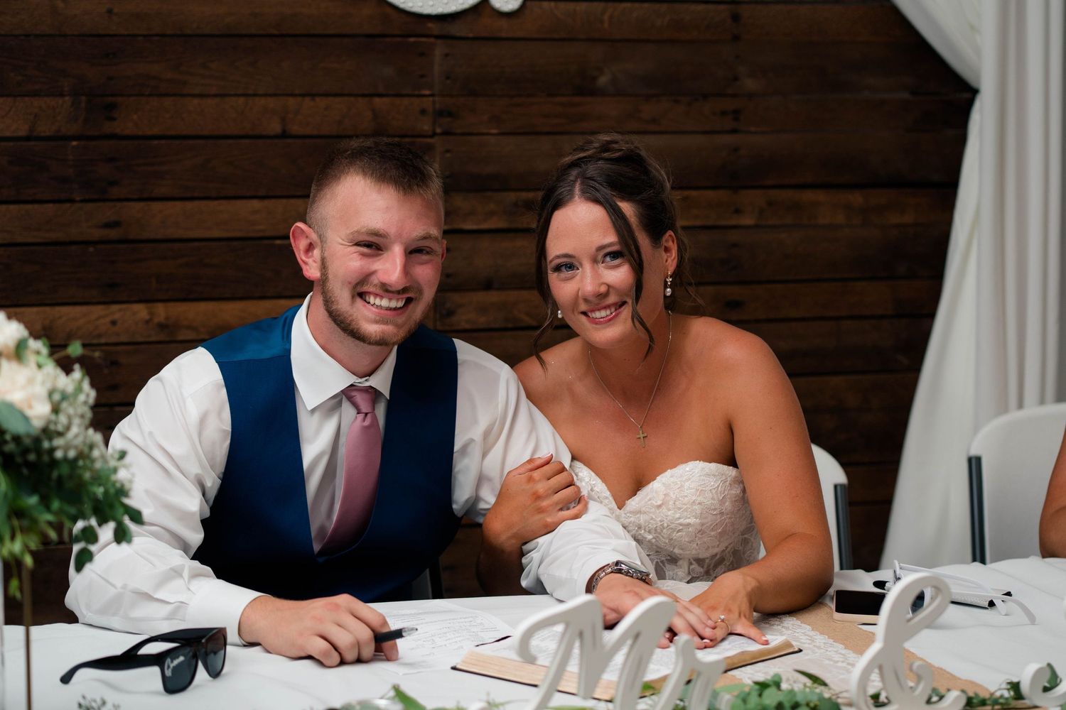 Smiling bride and groom sit together at sweetheart table with sunglasses and greenery decor at reception.