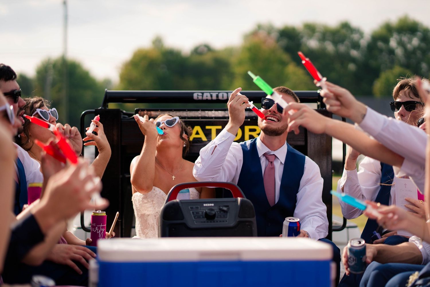 Bride and groom celebrate with guests on decorated wagon ride, wearing fun sunglasses and holding glow sticks outdoors.