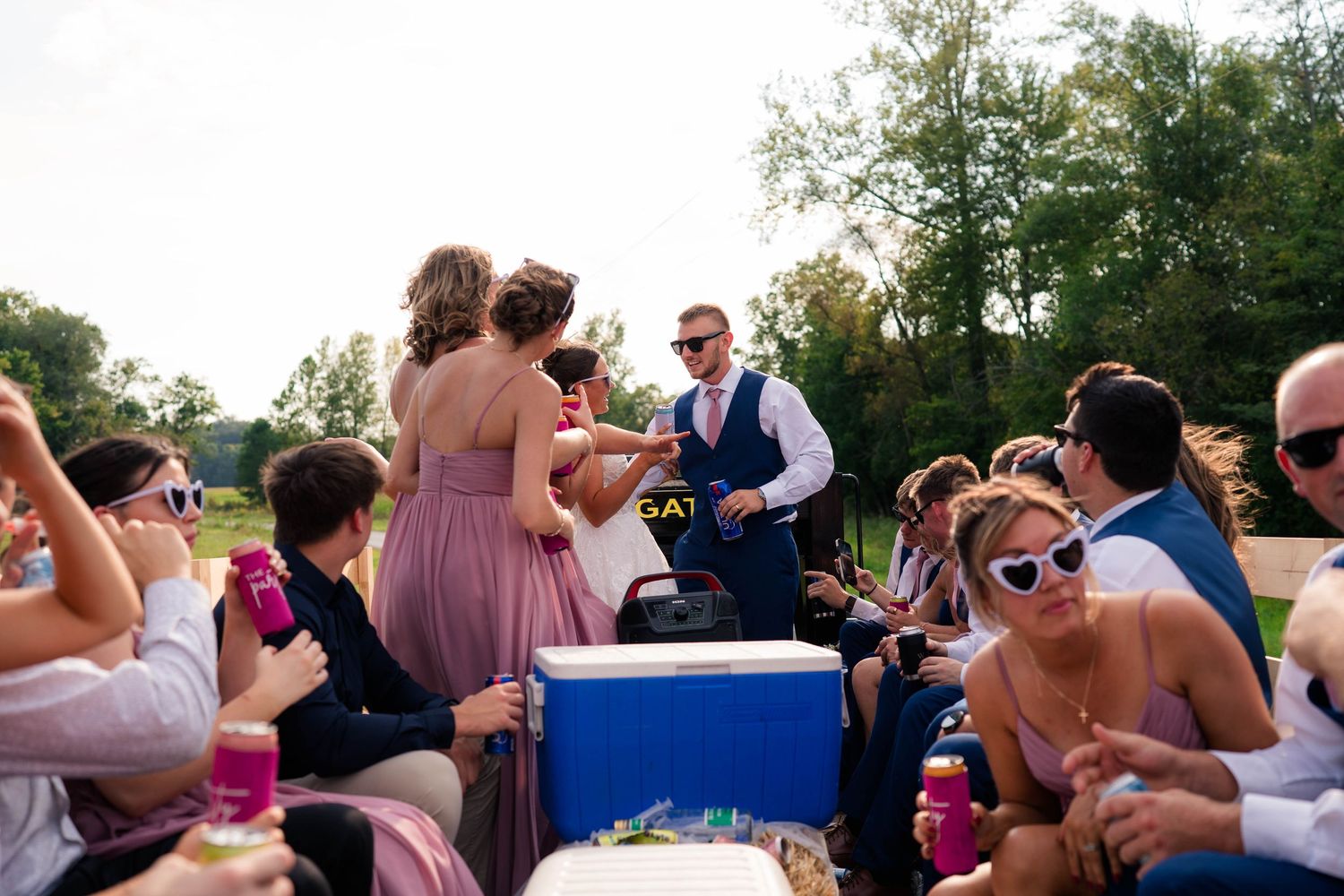 Groom on tractor distributes drinks to wedding guests during outdoor reception celebration.