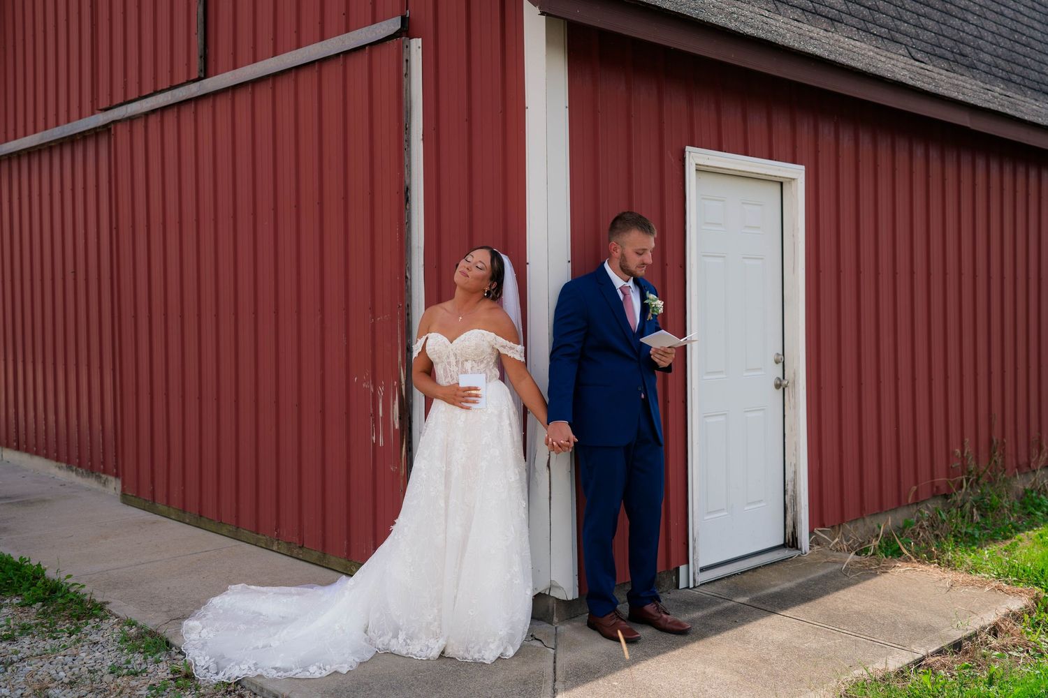 Bride laughing joyfully while groom reads vows during their first look beside a red barn.