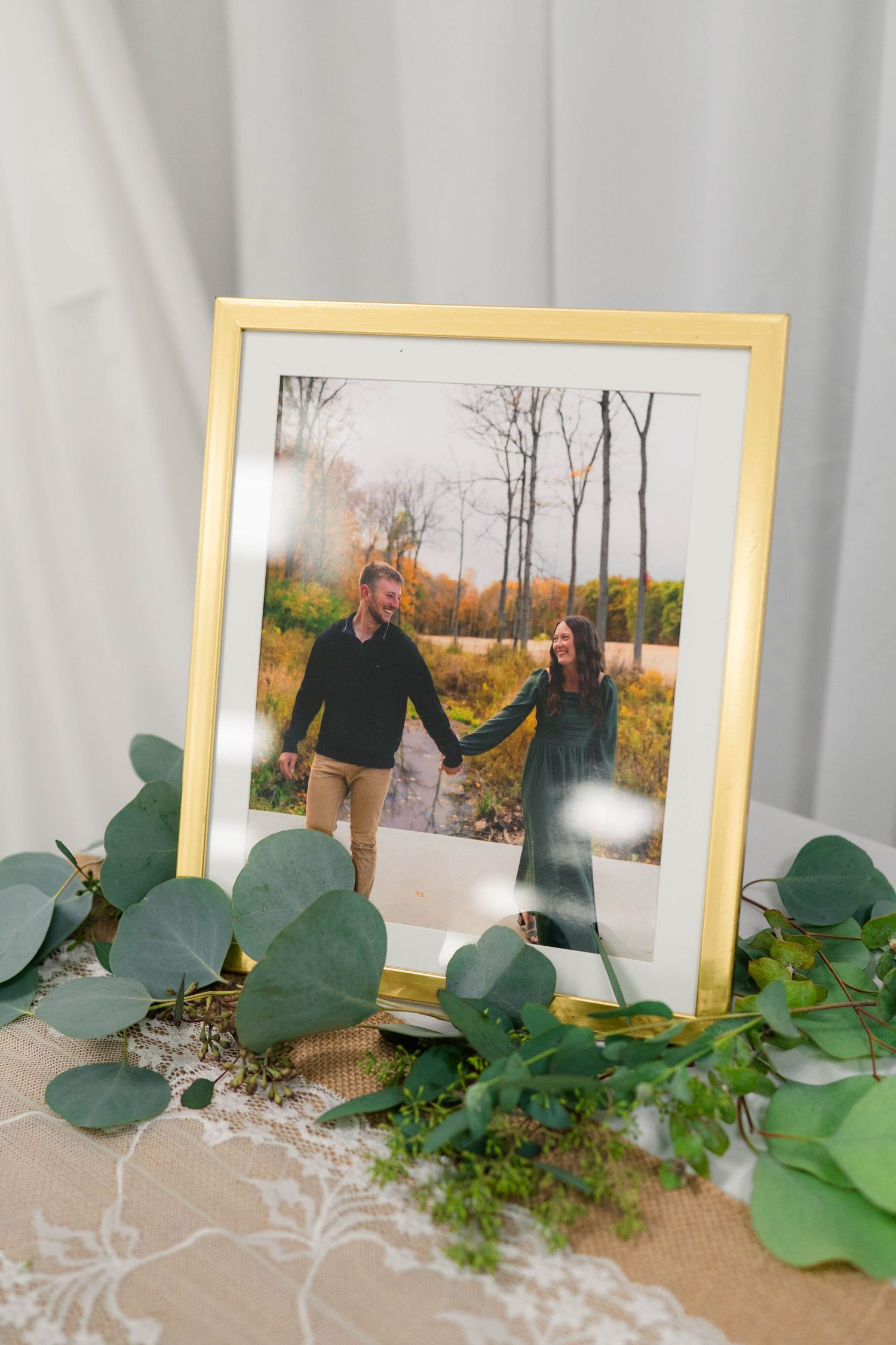 Gold framed engagement photo of couple holding hands in autumn field with eucalyptus.