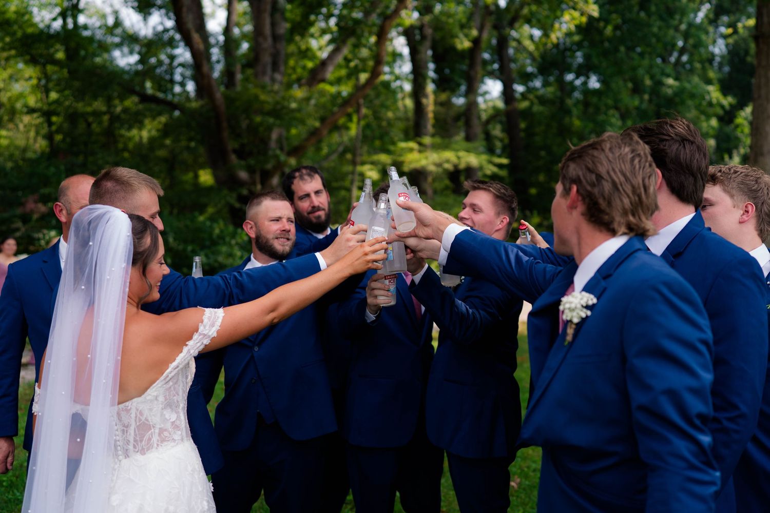 Bride and groomsmen in navy suits toasting drinks together outdoors at wedding celebration.