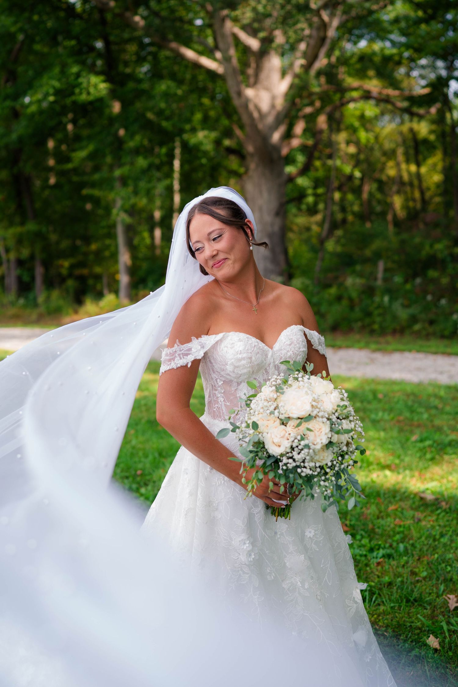 Radiant bride in off-shoulder lace gown holds white bouquet as her veil flows in the breeze.