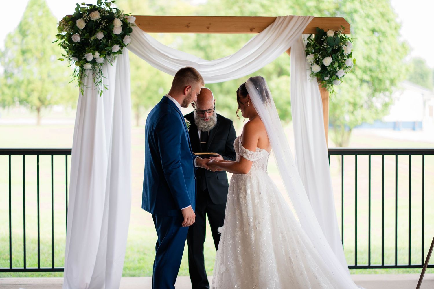 Bride and groom exchanging vows under a floral arch with white draping at an outdoor wedding.