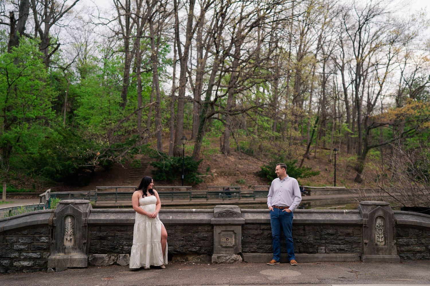 Pregnant woman and man pose apart at stone terrace with lush trees in park engagement photos.
